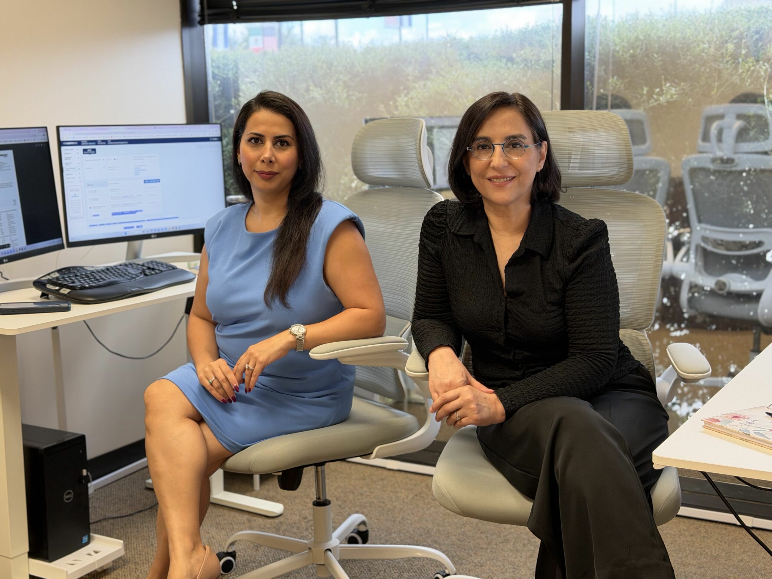 Two women sitting in office chairs in front of computer monitors, smiling, with a window and cars outside in the background.