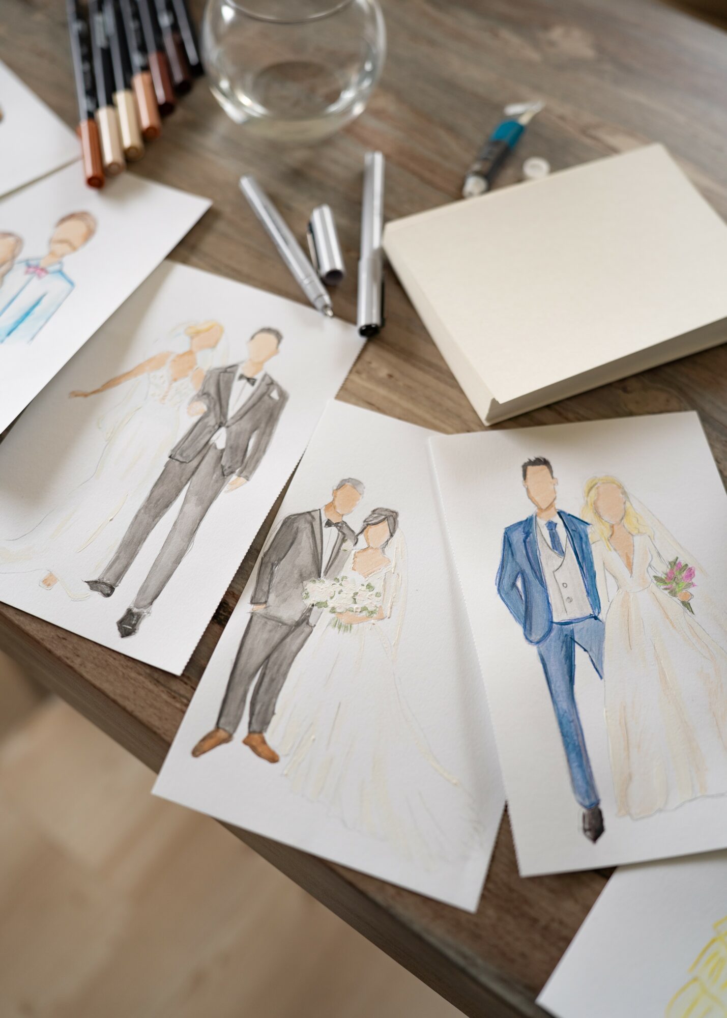 Fashion sketches of men in suits and women in dresses on a wooden table with pens, paper, and a glass.