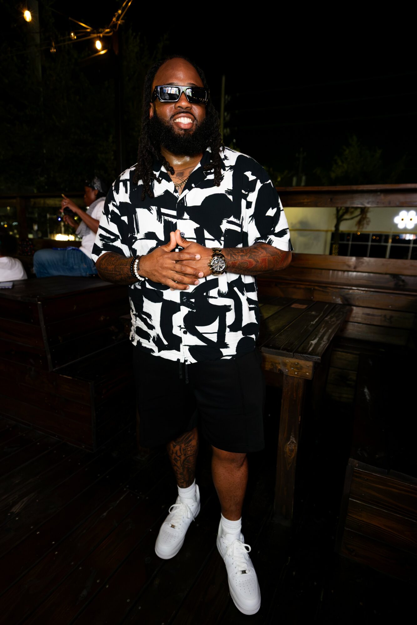 Man with sunglasses and beard wearing a black and white patterned shirt, black shorts, and white sneakers, standing outdoors at night.