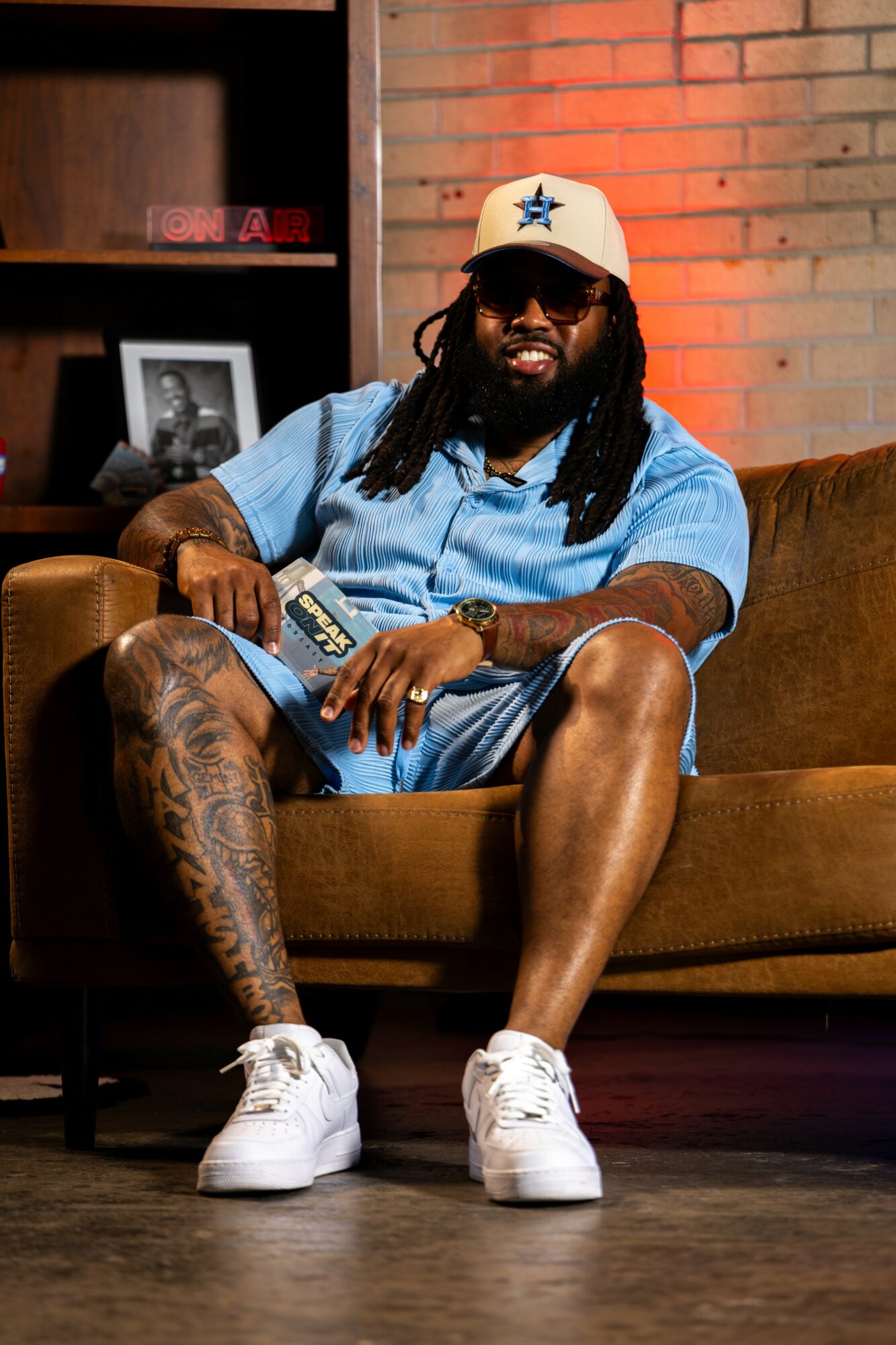 Man with dreadlocks, sunglasses, and a white cap with a star, sitting on a brown couch, holding a remote, in a room with brick wall.
