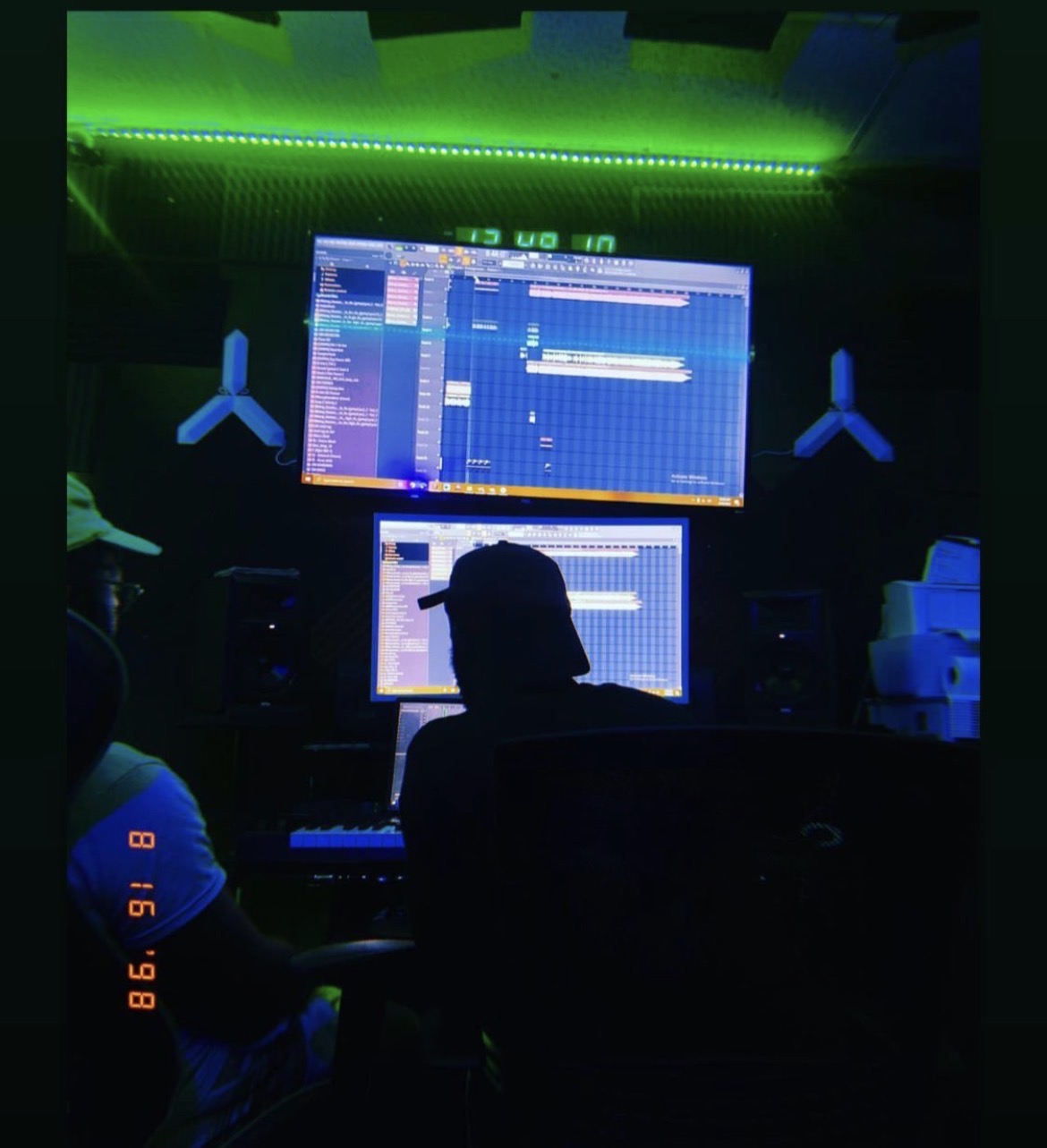 Person working on music production in a dark studio with two monitors and green LED lighting, silhouette visible.