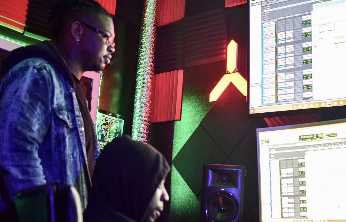 Two people in a music studio with computer screens and colorful lighting, one standing and one sitting, focused on screens.