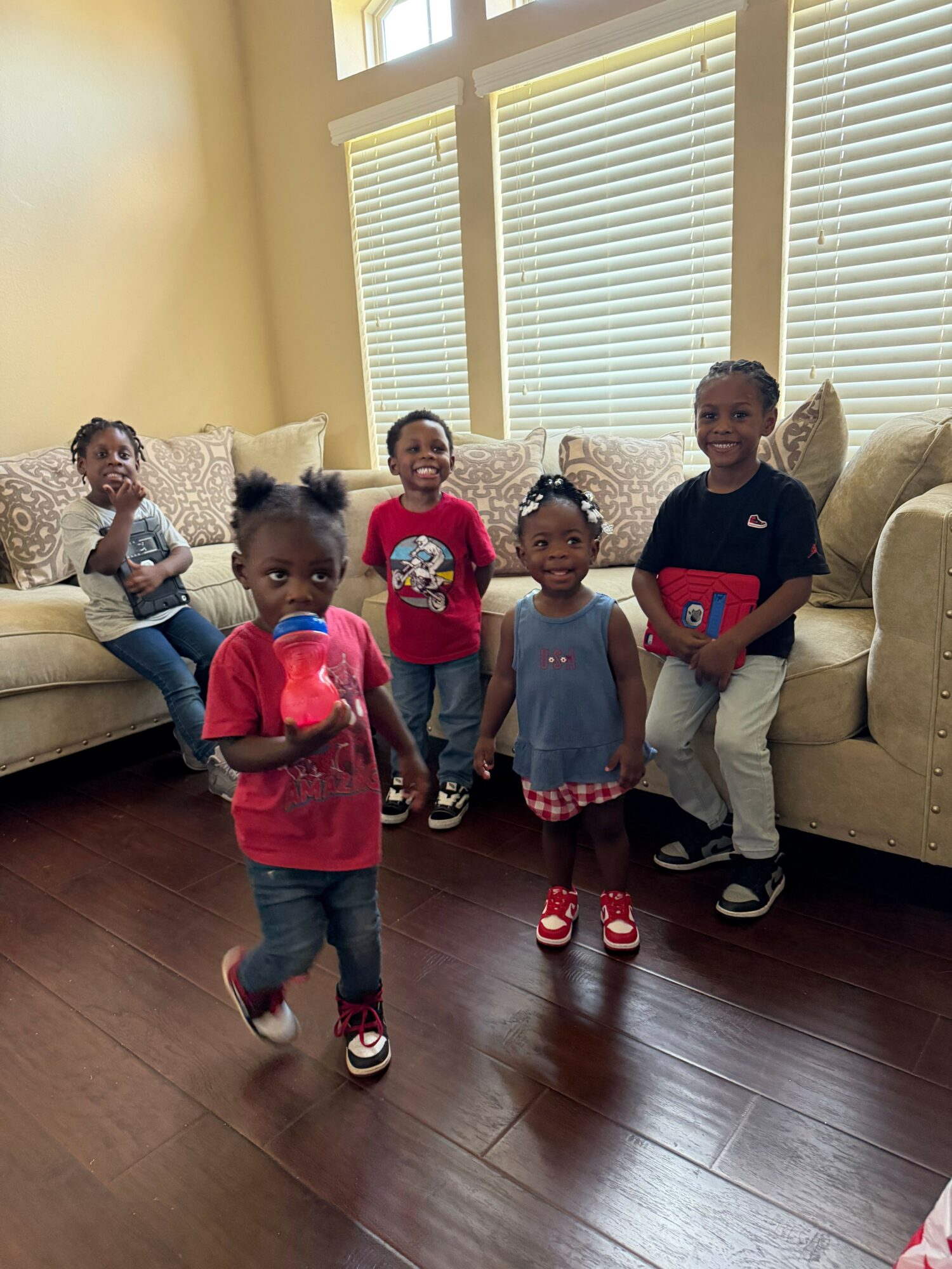 Four children and one adult standing in a living room with wooden floor and large window with blinds.