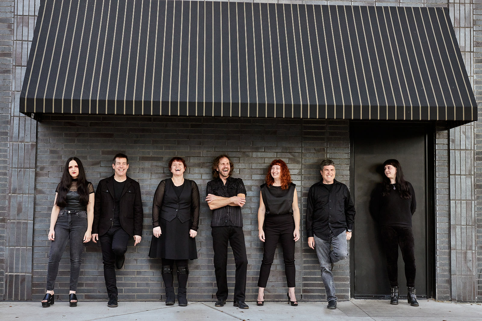 Seven people stand in front of a brick wall with a striped awning above them, facing forward.