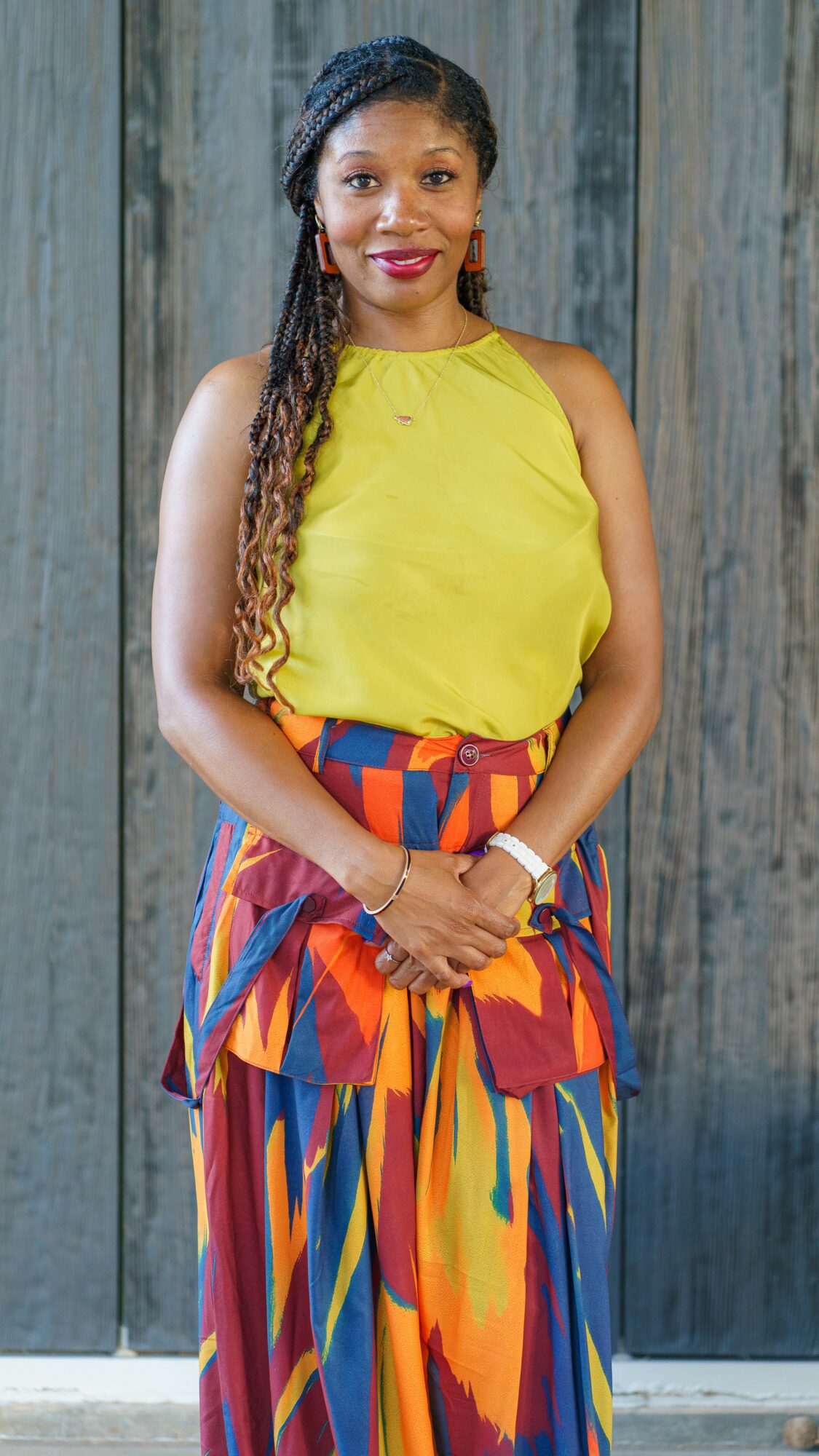 Woman with long braided hair wearing a yellow top and colorful skirt, standing in front of a wooden background.