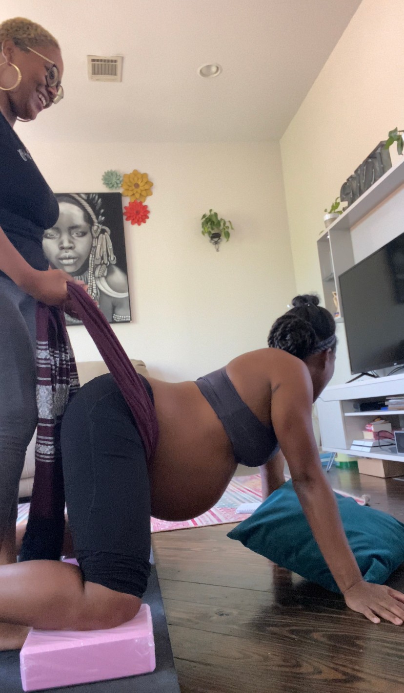 Pregnant woman on hands and knees on floor, supported by another woman standing nearby, in a living room.