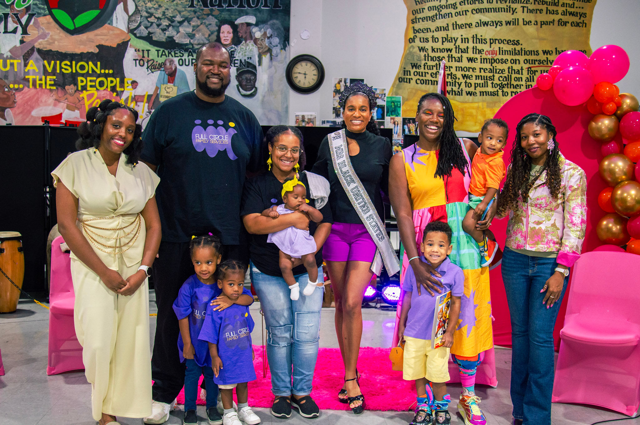 Group of adults and children smiling indoors with colorful decorations and a pink chair, celebrating an event.