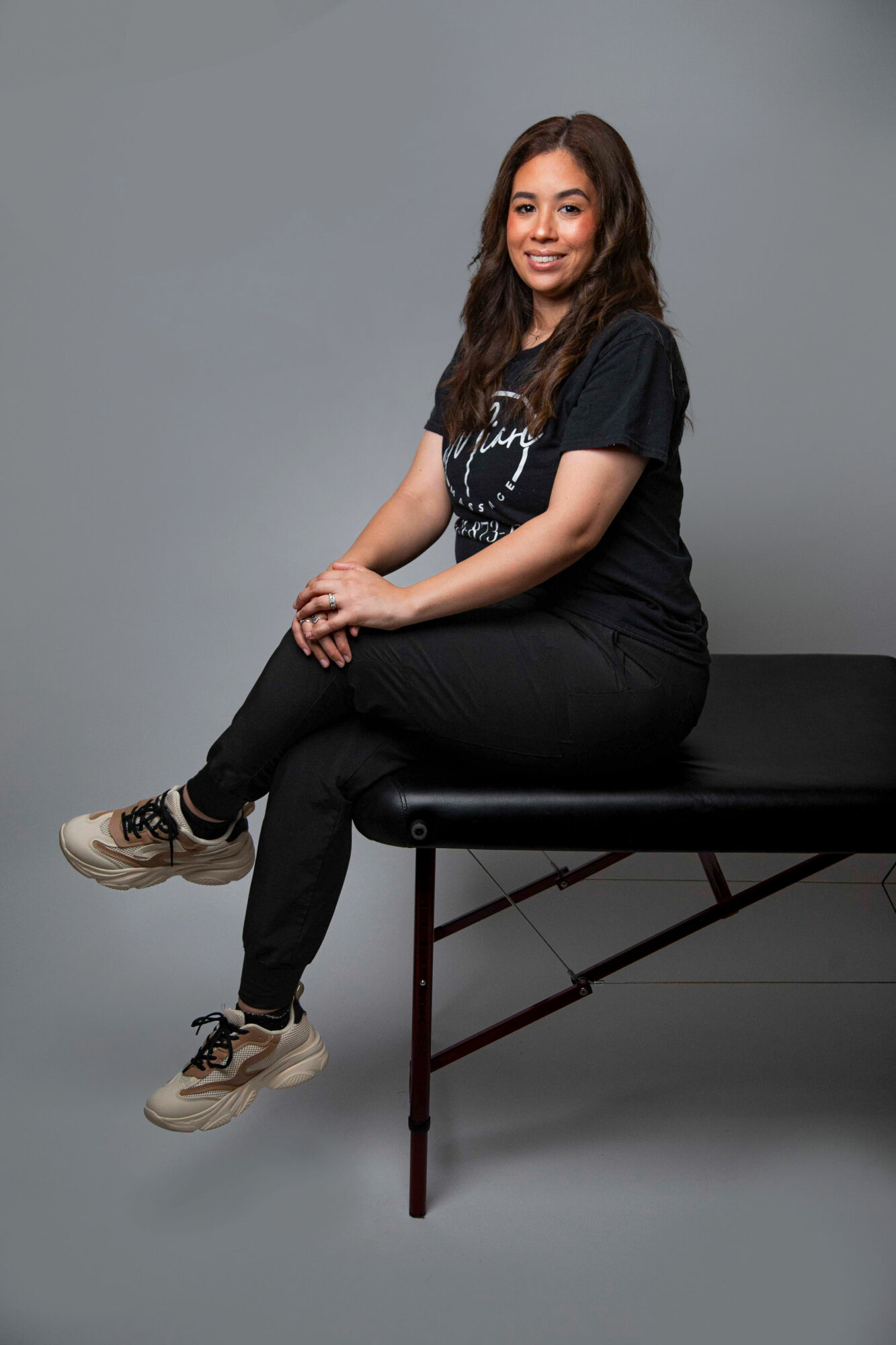 Young woman sitting on a black bench, smiling, wearing a black t-shirt, black pants, and sneakers, against a gray background.