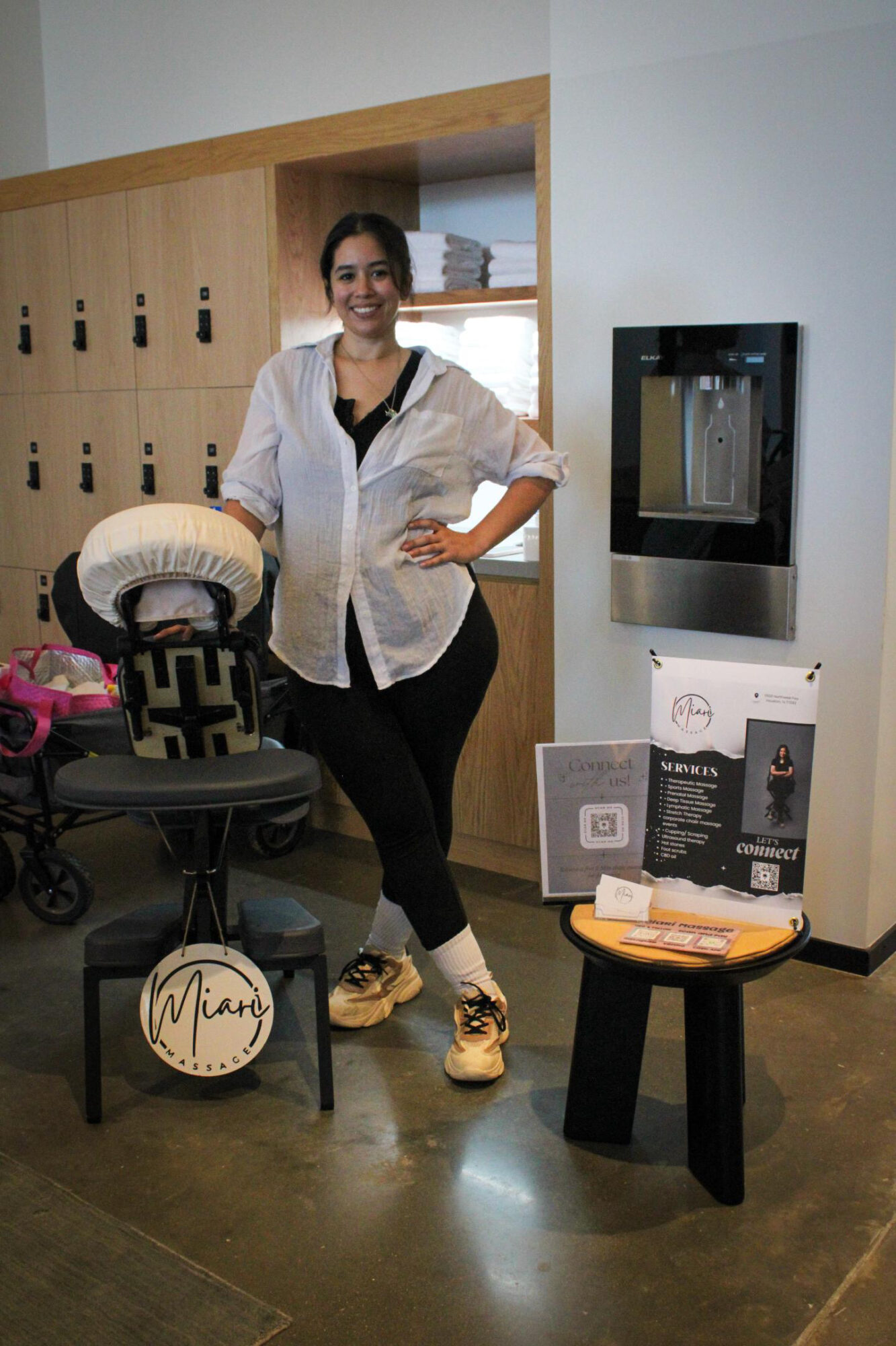 Woman standing indoors next to a stroller and a table with promotional materials, smiling, with lockers and a vending machine behind her.