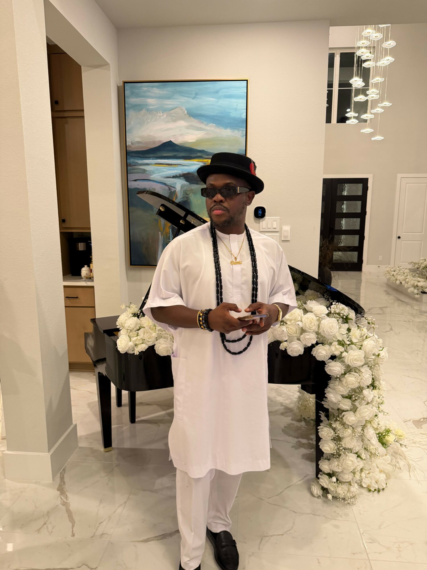 Person dressed in white with a black hat, holding a camera, standing in a decorated room with flowers and artwork.