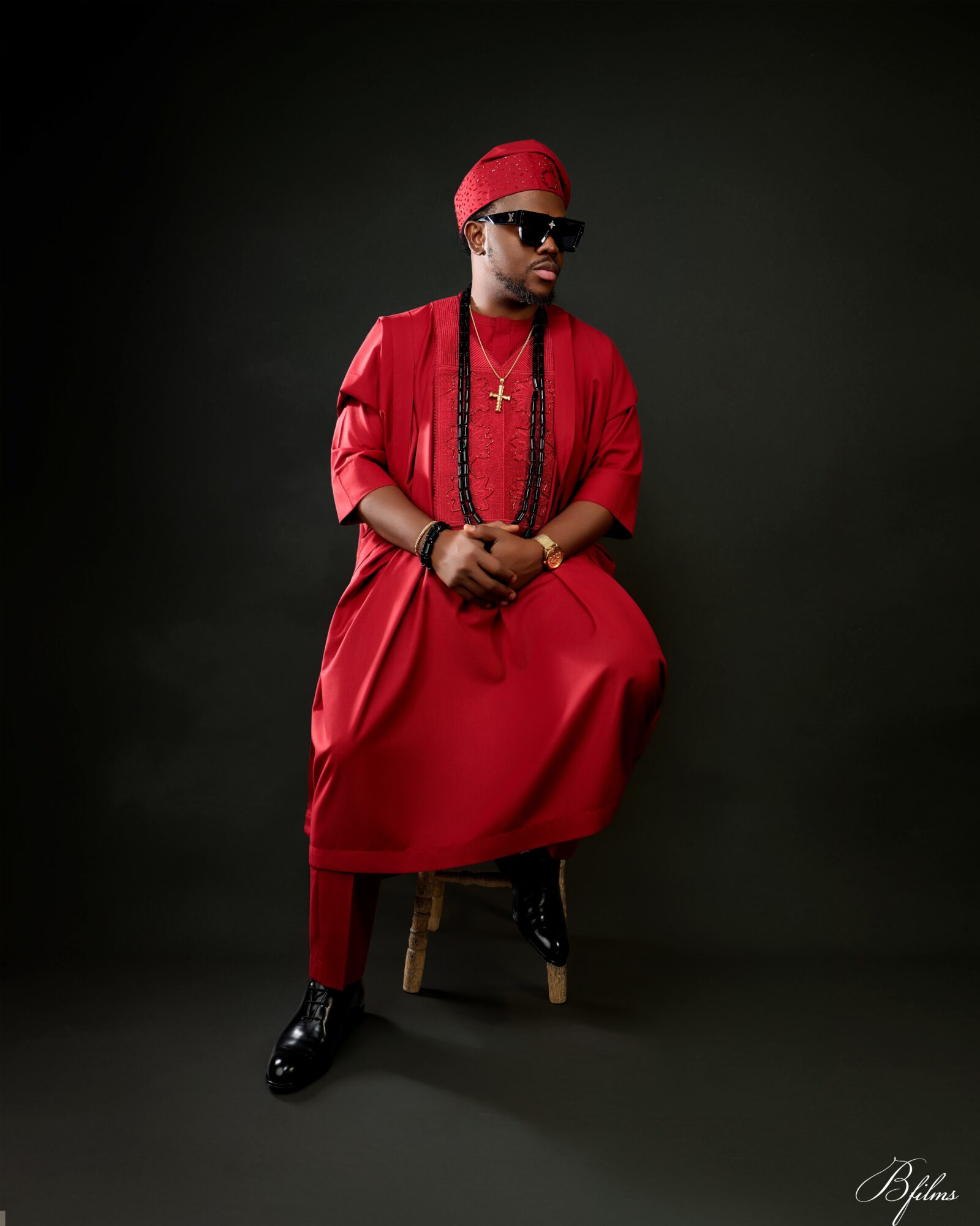 Person dressed in red sitting on a stool, wearing sunglasses, headscarf, and jewelry, against a dark background.