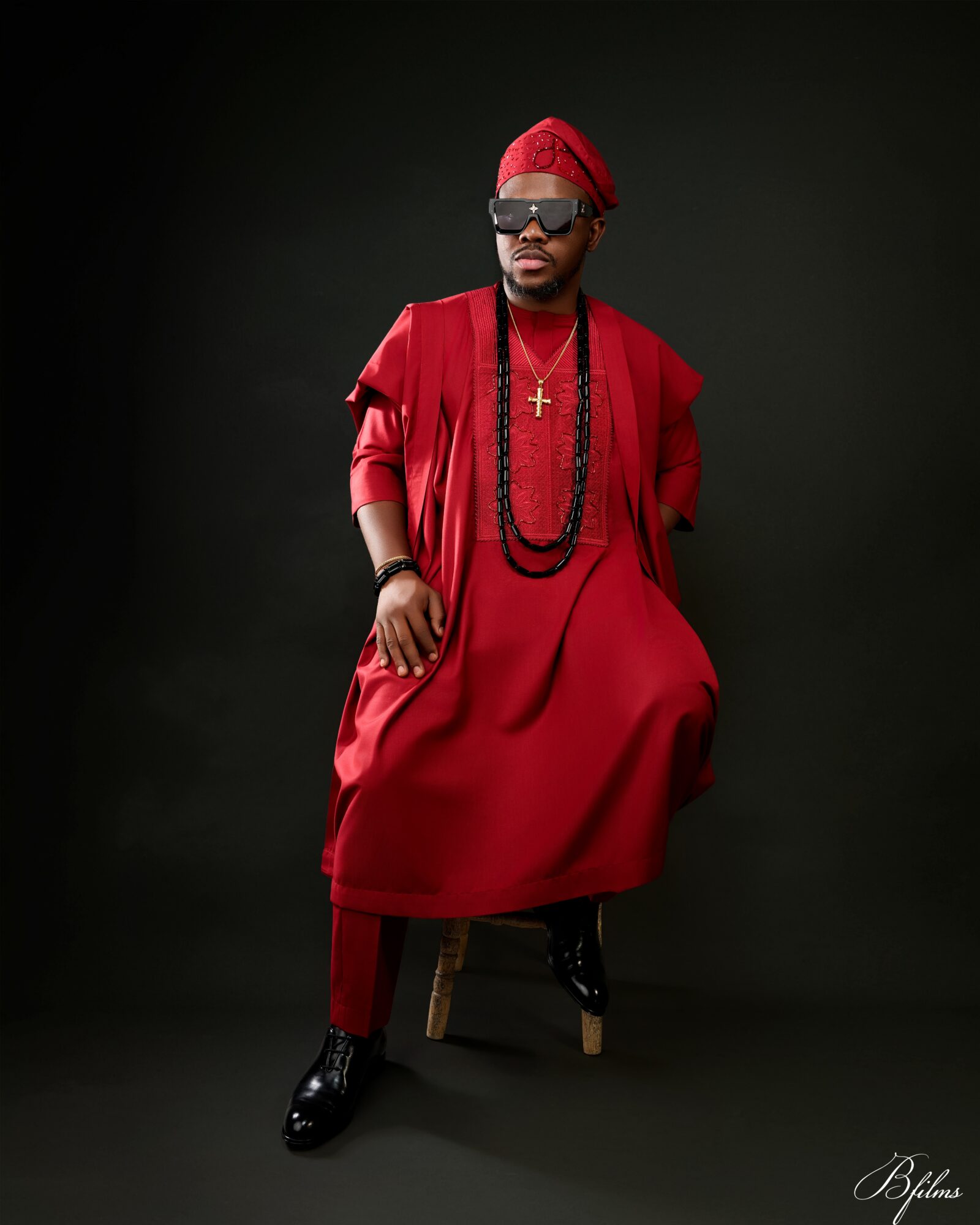 Man dressed in red traditional attire with accessories, sitting on a stool against a dark background.