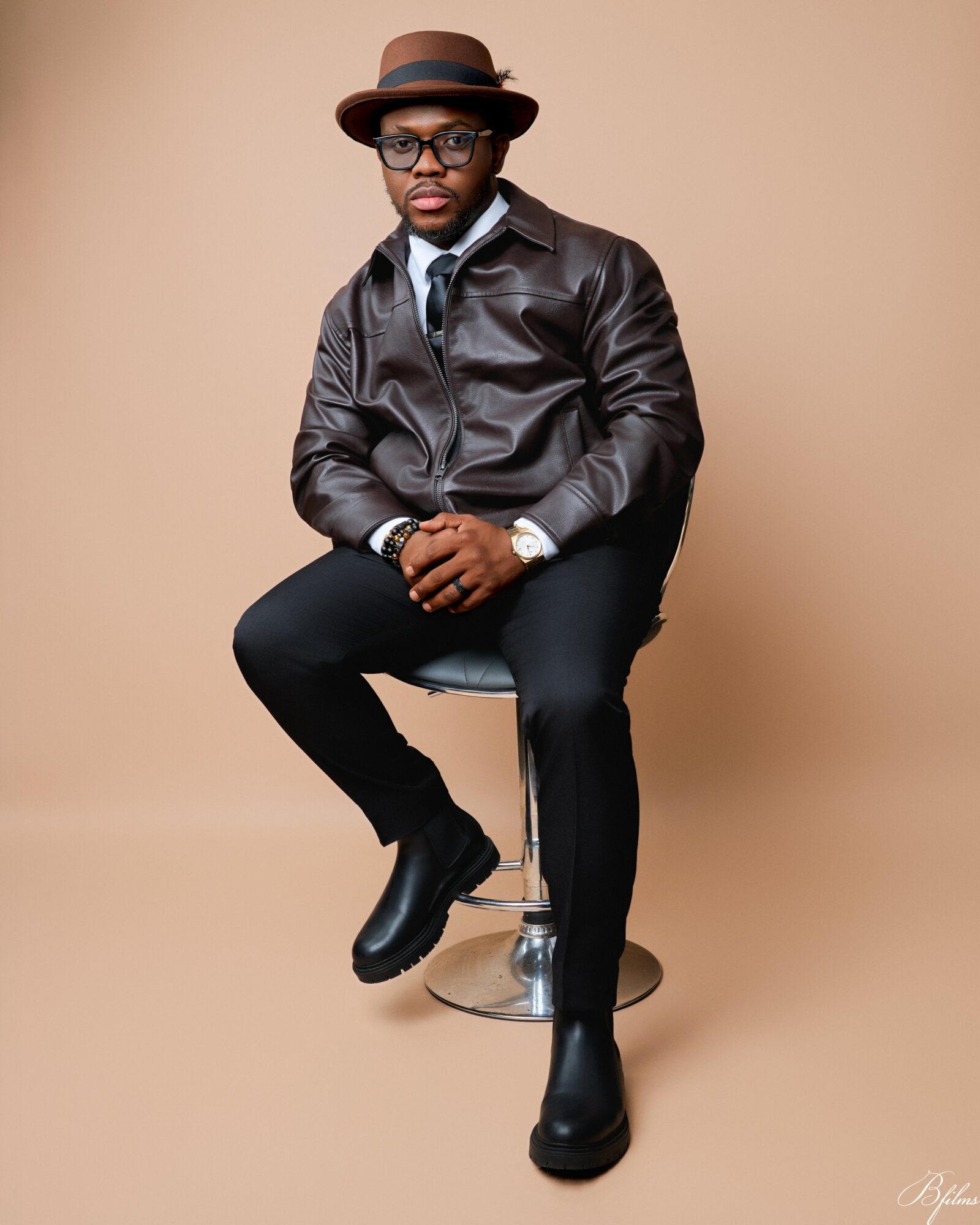 Person sitting on a stool wearing a leather jacket, hat, glasses, and black boots against a beige background.