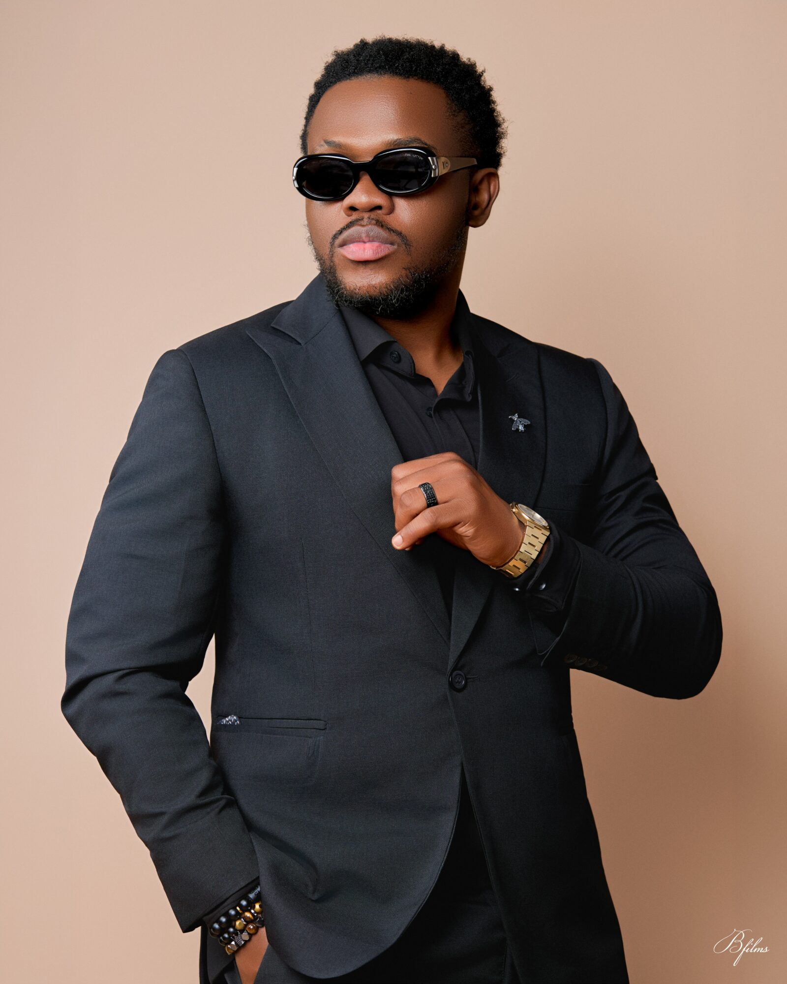 Man in black suit and sunglasses adjusts his jacket against a beige background.