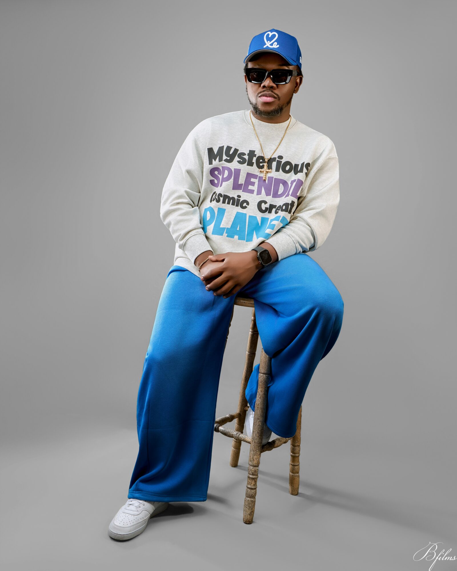 Man sitting on a wooden stool, wearing a blue cap, sunglasses, a white sweatshirt with colorful text, and blue pants.