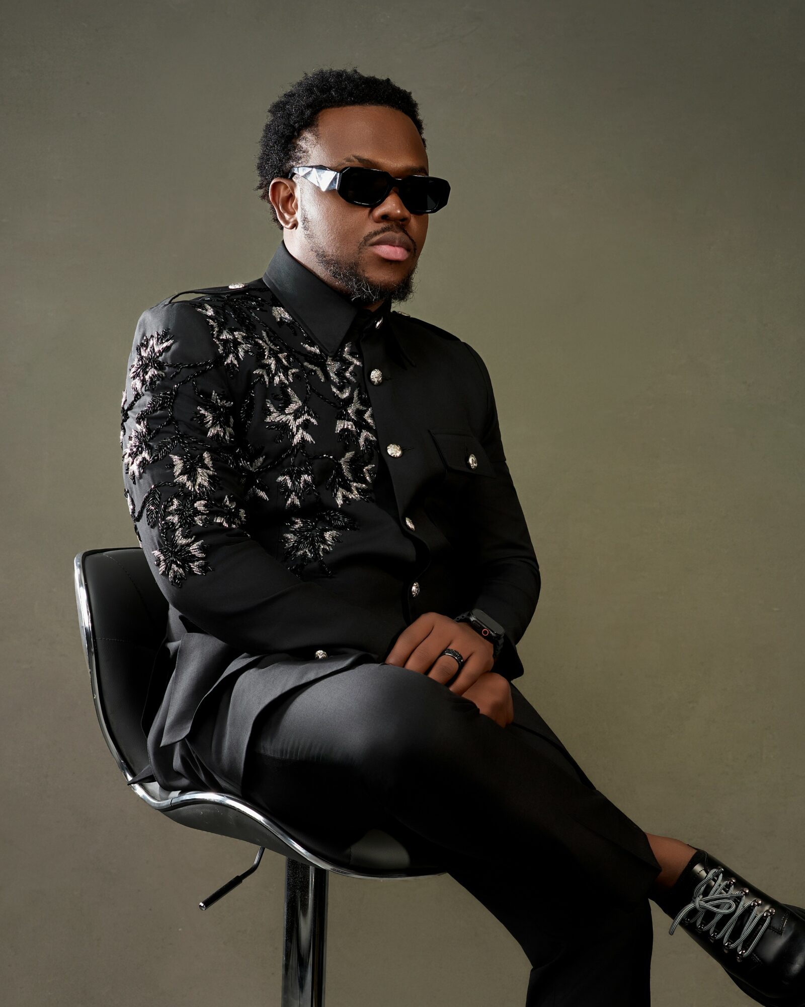 Man with sunglasses sitting on a chair, wearing a black and patterned jacket, black pants, and sneakers, against a plain background.