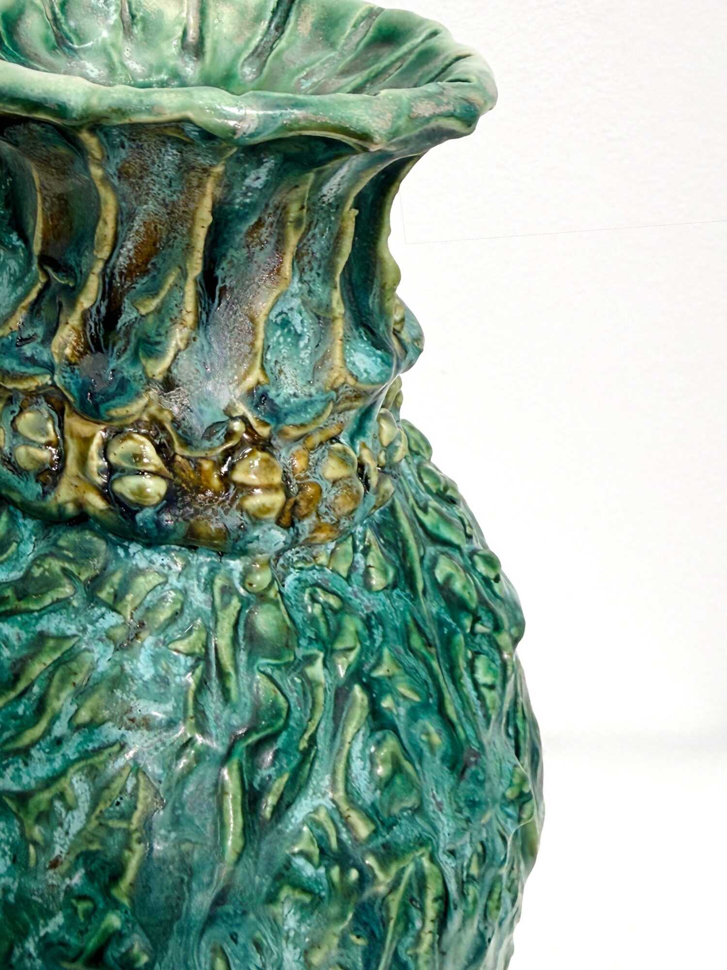 Close-up of a textured, colorful ceramic vase with green, blue, and gold tones against a plain white background.