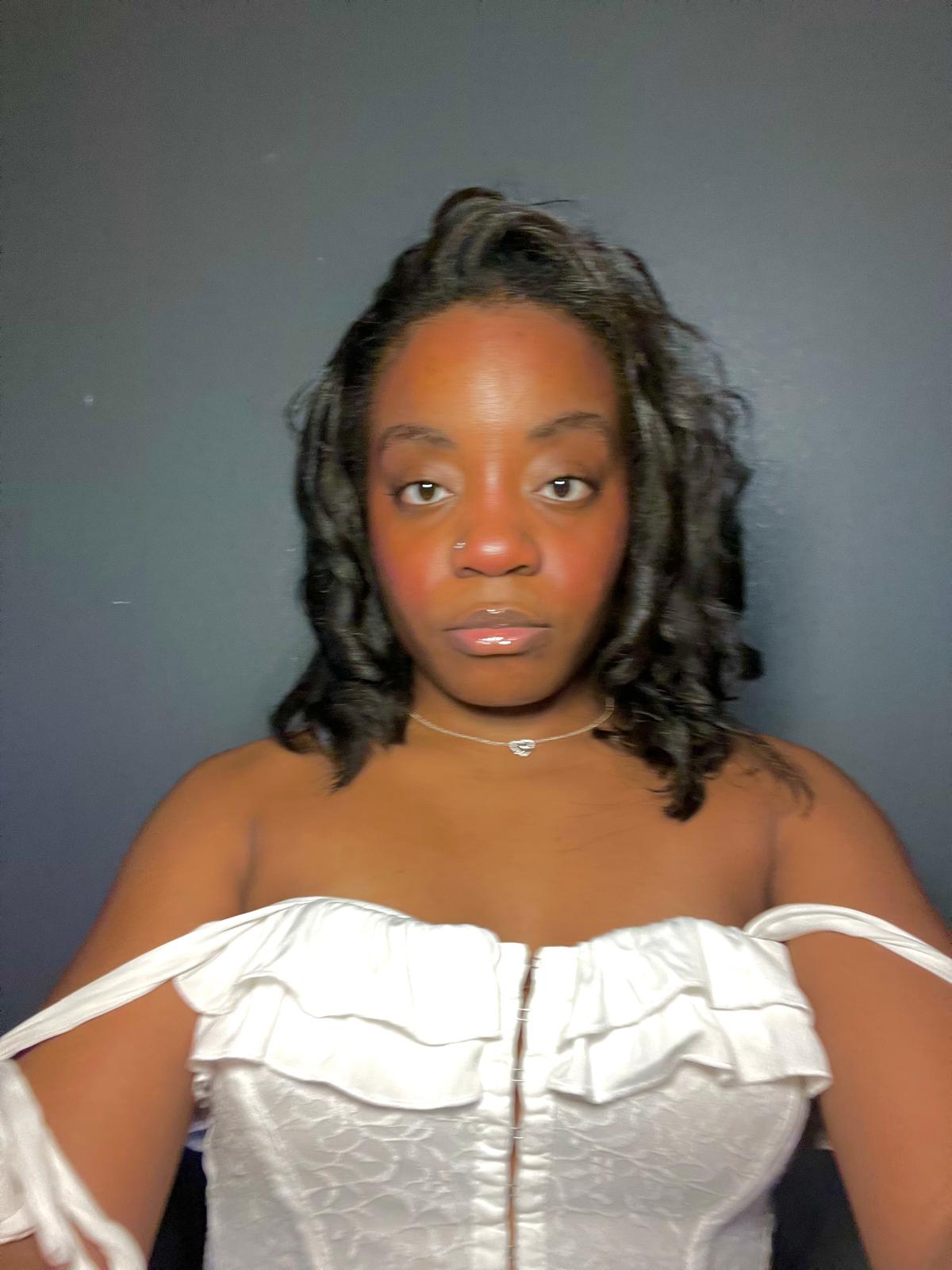 Young woman with shoulder-length curly hair, wearing an off-shoulder white top, looking directly at the camera against a dark background.