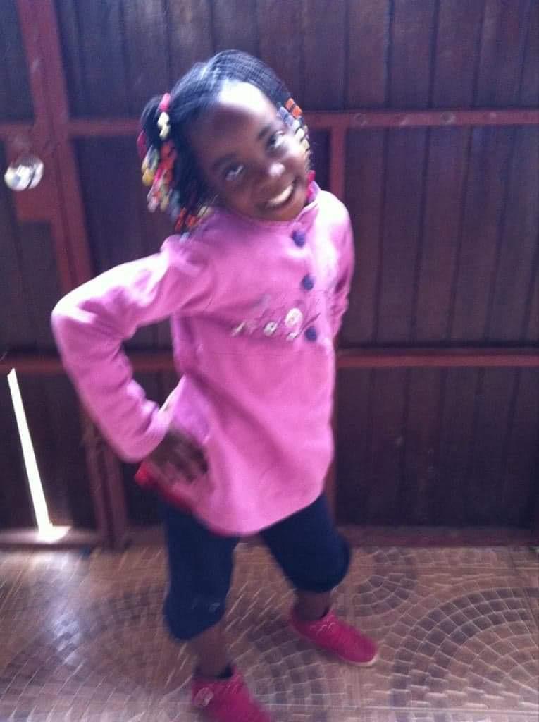 Young girl with braided hair and colorful beads, smiling, wearing a pink coat, black pants, and red shoes, standing indoors.