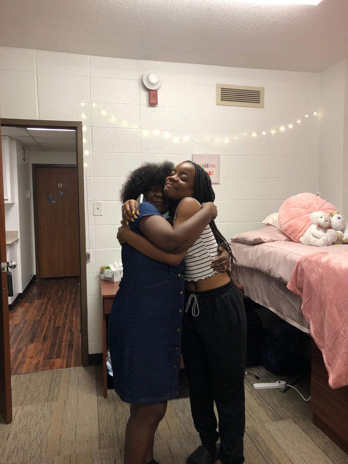 Two people hugging in a room with a bed, pink blanket, and stuffed animals, wall string lights, and a door in the background.