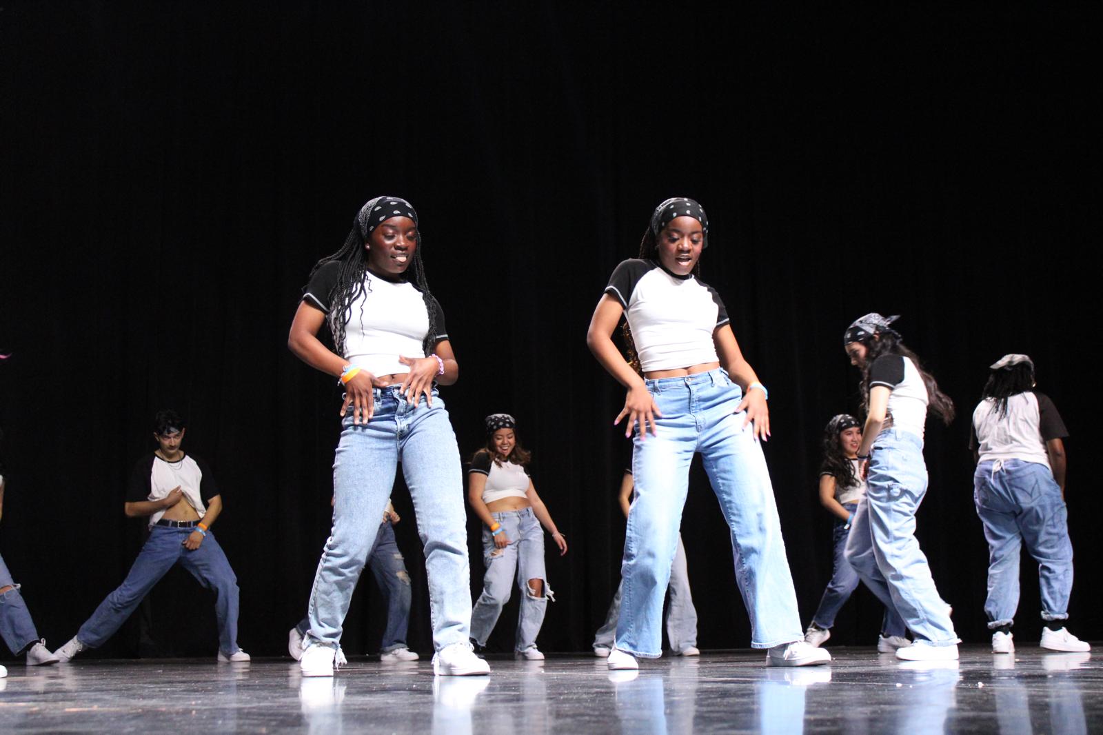 Group of young women dancing on stage, wearing casual outfits with jeans and t-shirts, performing synchronized dance moves.