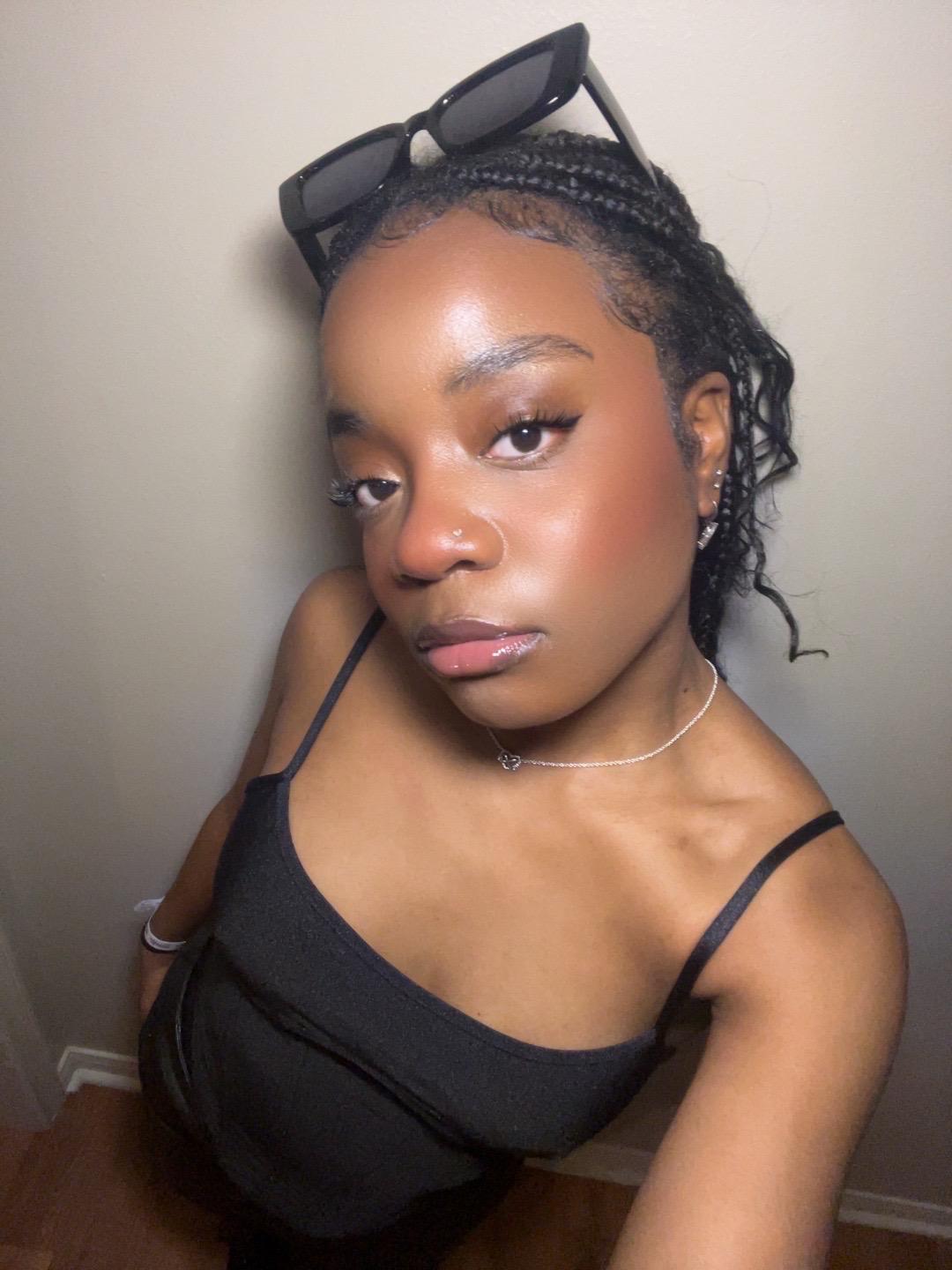 Young woman with braided hair, sunglasses on head, wearing a black top, looking at camera with neutral expression.