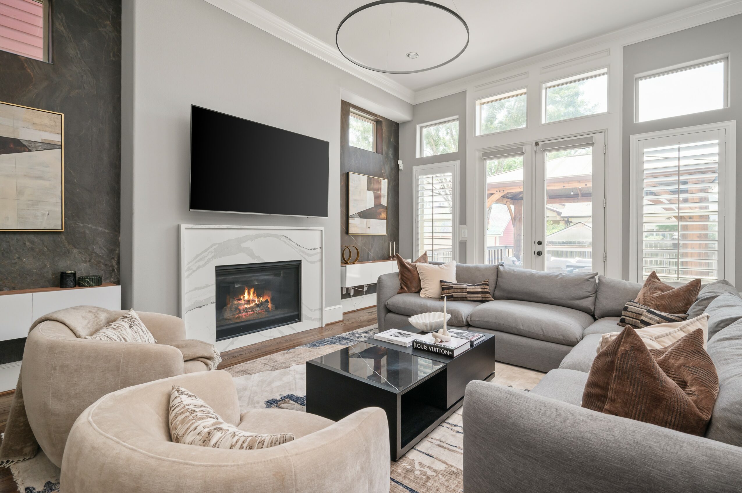 Living room with large windows, a fireplace, a wall-mounted TV, and beige and gray furniture, including a coffee table.