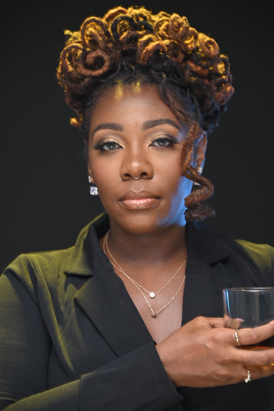 Woman with styled curly hair, wearing a black blazer and jewelry, looking at the camera against a dark background.