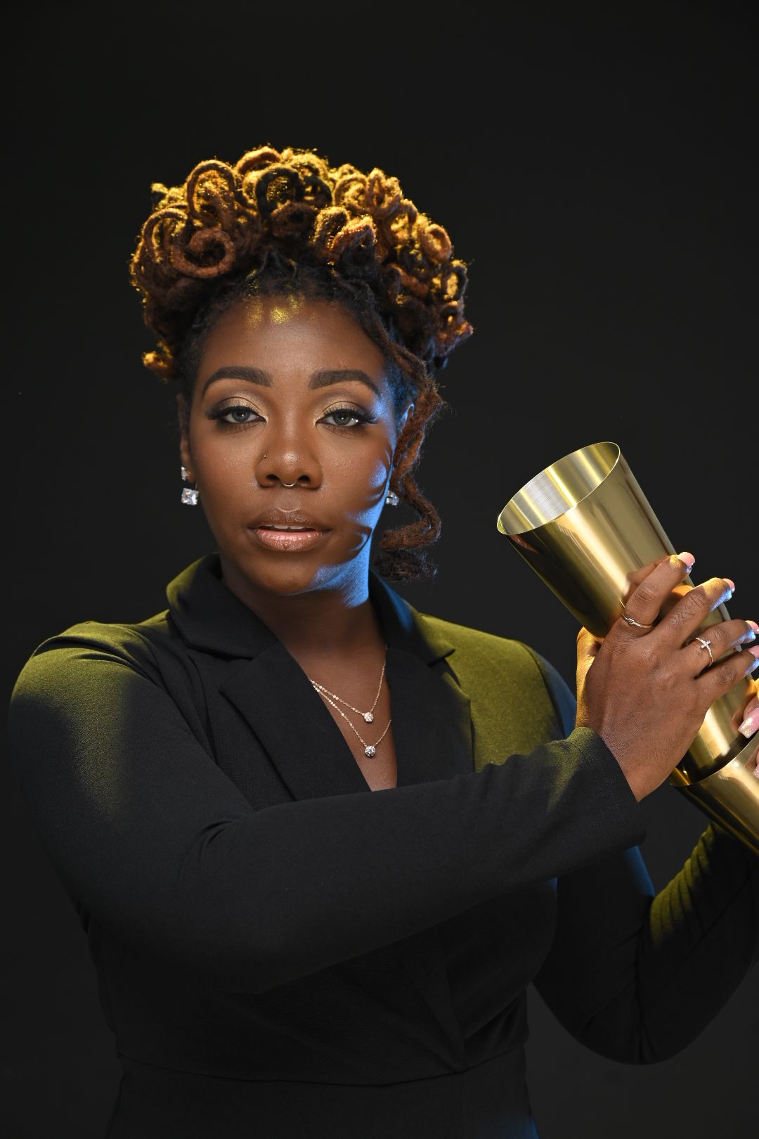 Woman with curly hair holding a gold-colored trophy, wearing a black blazer, against a dark background.