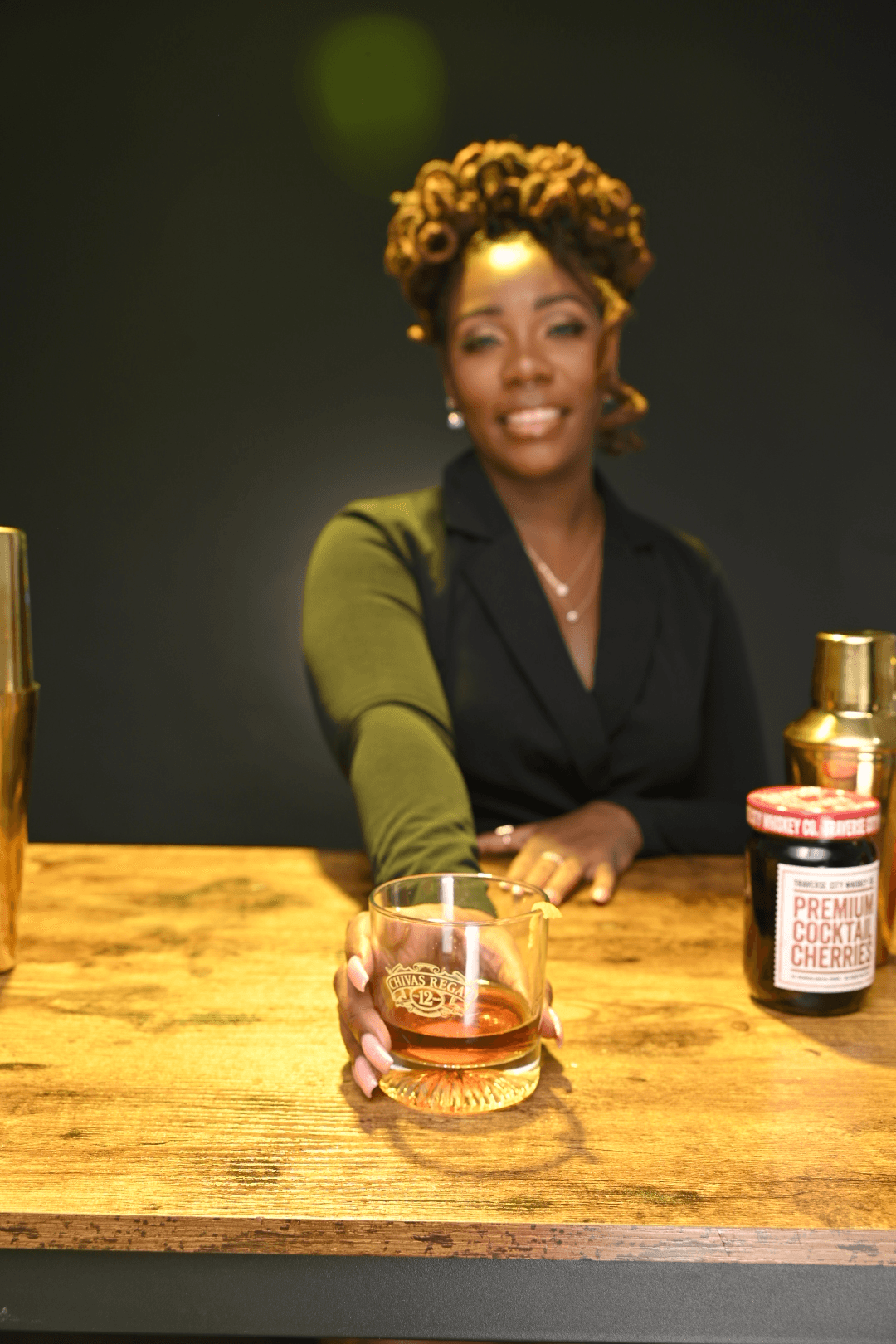Woman with curly hair smiling, sitting at a wooden table with a drink and jars, dark background.