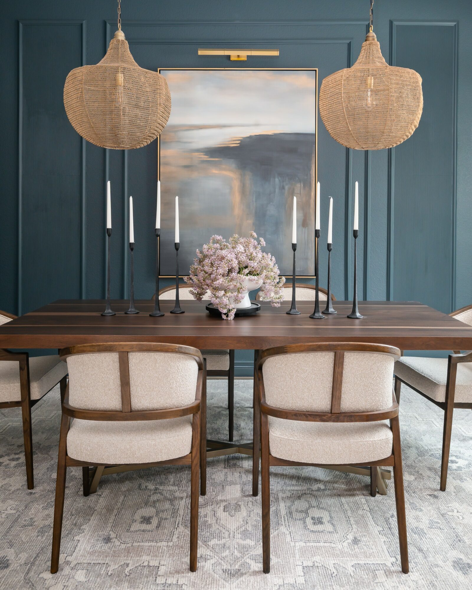 Dining table with six chairs, two hanging pendant lights, a mirror, and a flower arrangement, against a blue wall.