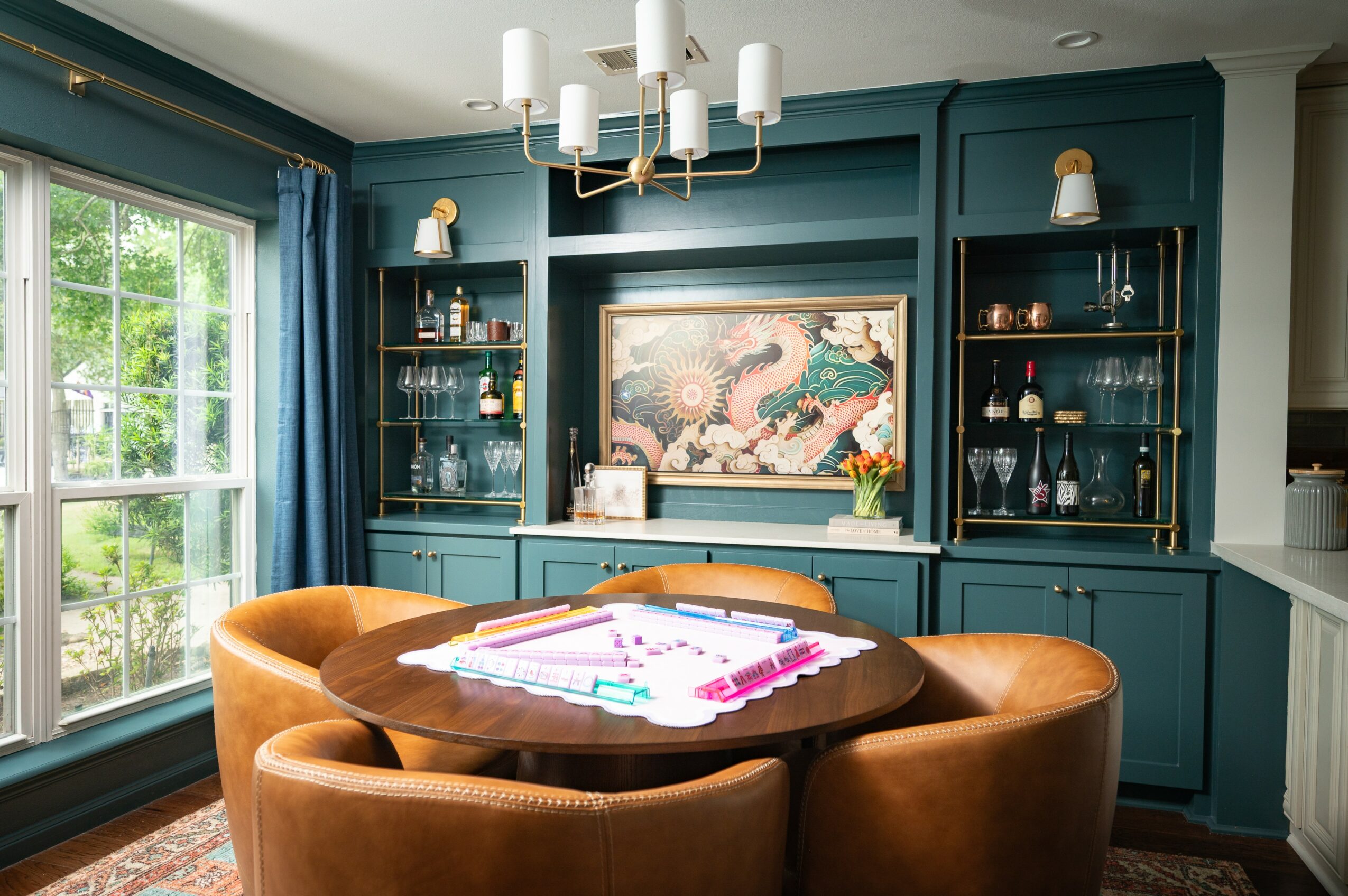 Dining room with teal wall, large window, round table with four brown chairs, artwork, and glass shelves with bottles and decor.