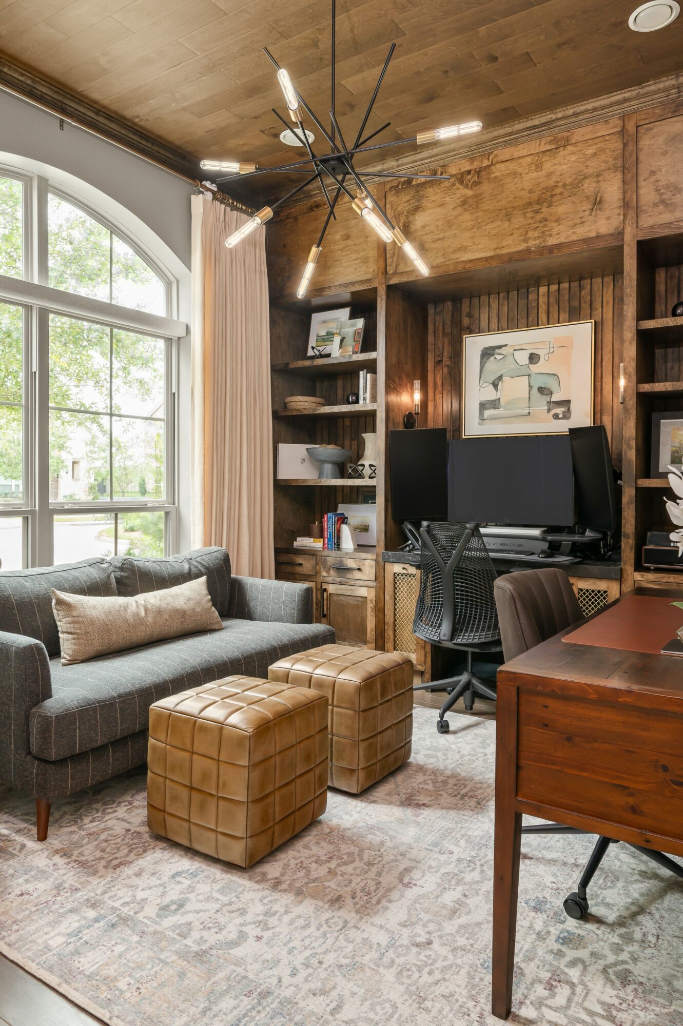 Living room with large window, gray sofa, two tan ottomans, wooden desk, bookshelf, and a black computer monitor.