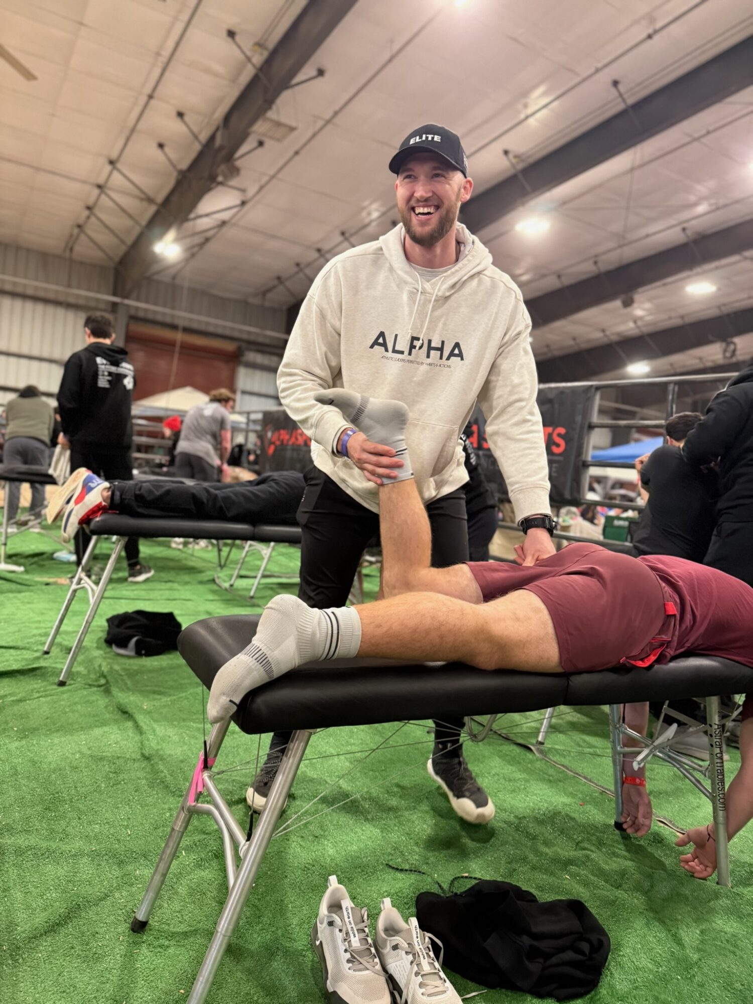 Man smiling and holding a water bottle stands next to a person lying on a massage table inside an indoor sports facility.