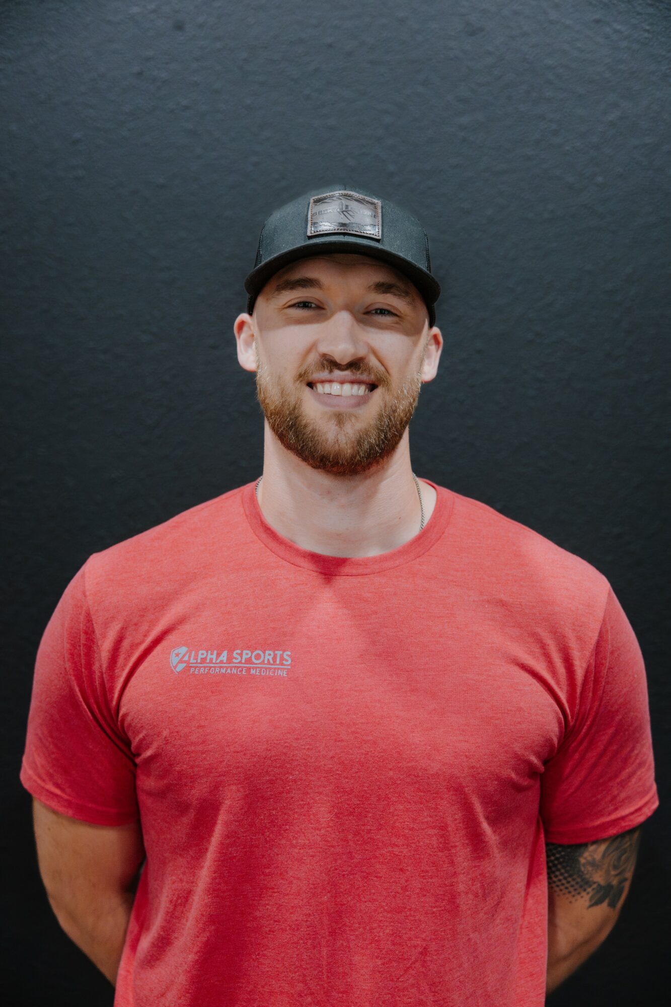 Smiling man with a beard wearing a black cap and red shirt stands against a dark background.