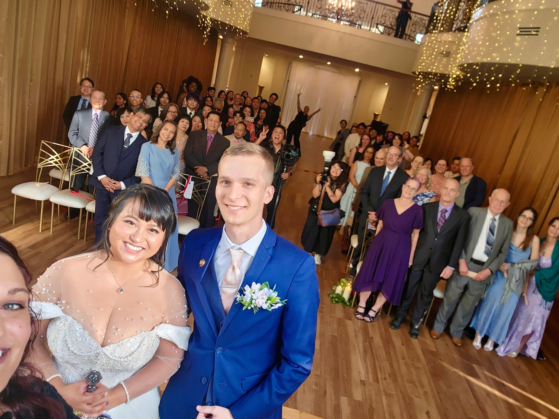 A group of people at a formal event, with two smiling individuals in the foreground, one in a blue suit and the other in a white dress, in a decorated hall.