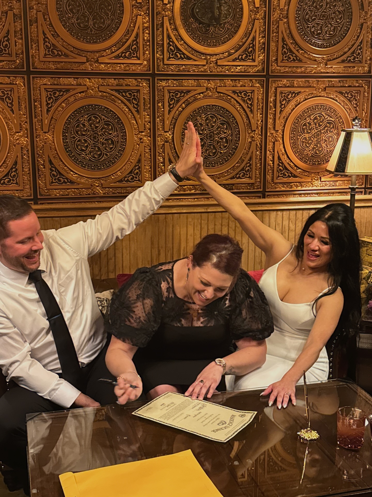 Three people celebrating with a certificate, high-fiving, in a decorated room with ornate ceiling tiles and a lamp. Word count: 20