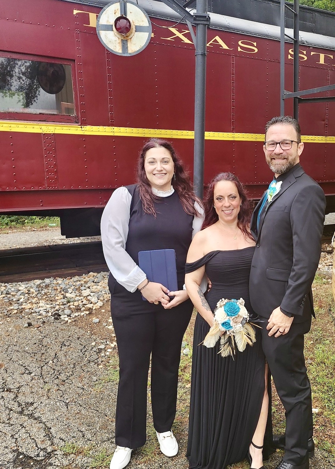 Three people standing outdoors in front of a red train car, smiling, with one woman holding a bouquet of flowers.