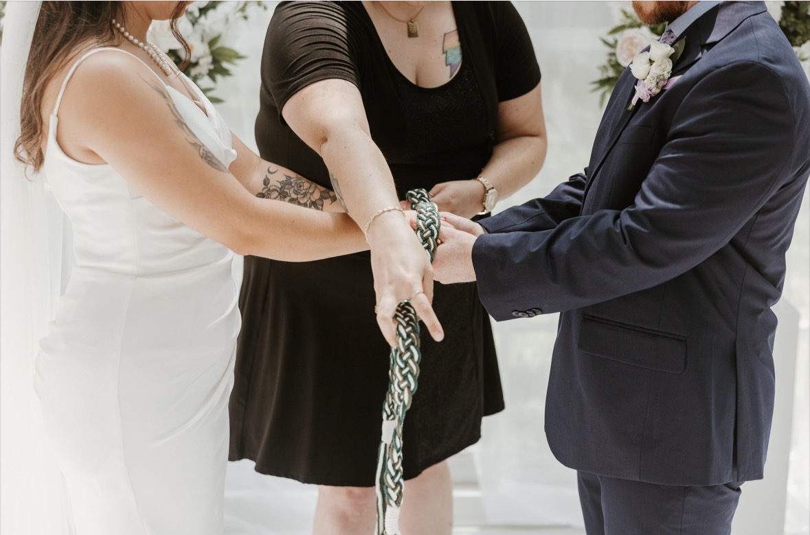 People holding hands during a wedding ceremony, with a woman in a white dress, another in black, and a man in a suit.
