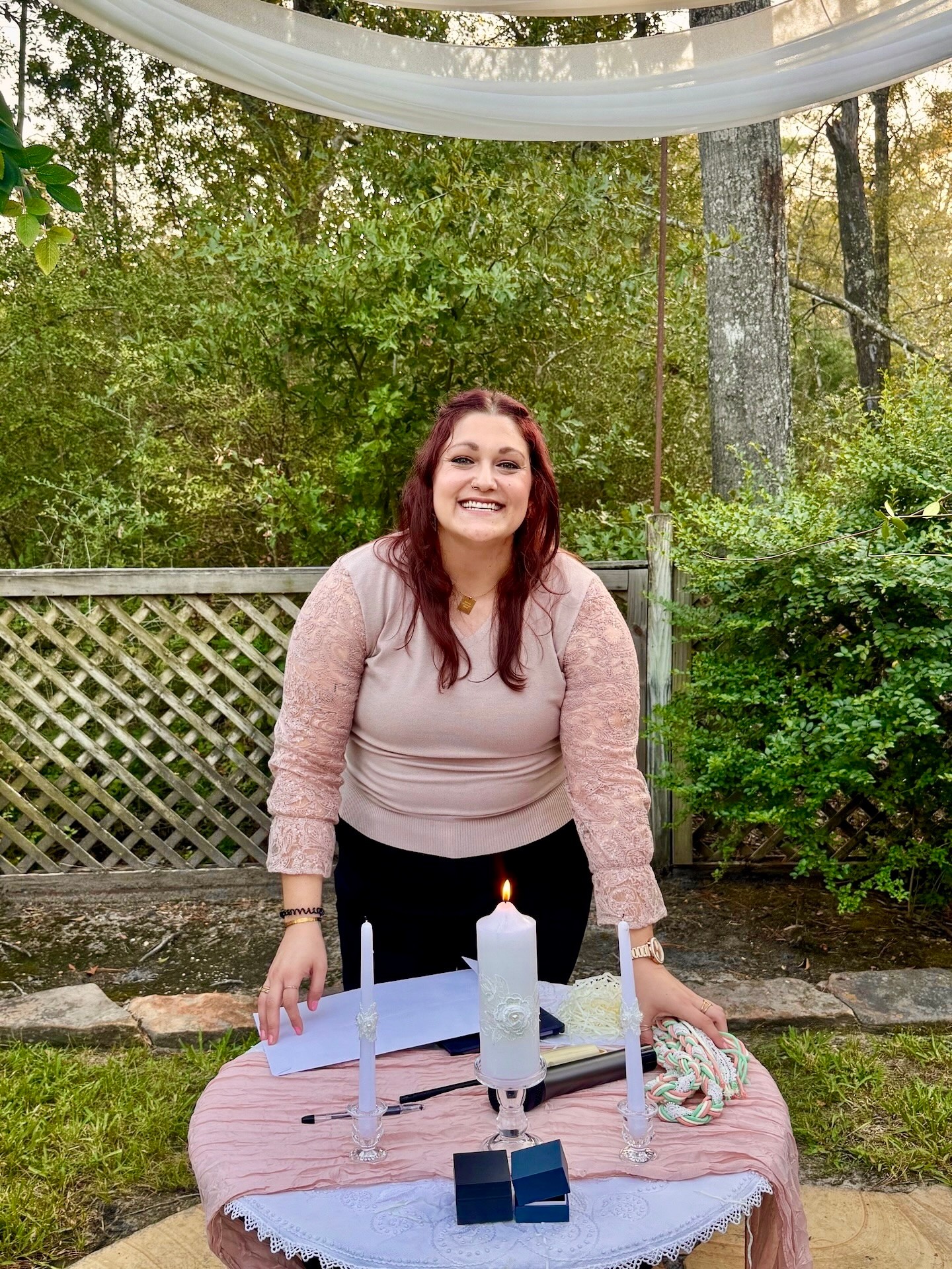 Woman with red hair smiling outdoors behind a decorated table with candles and a cake, surrounded by trees and greenery.