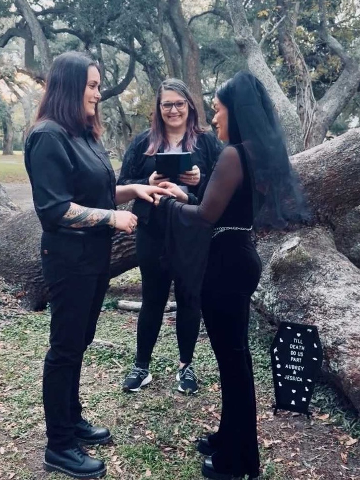Three women standing outdoors, one holding a small object, trees and rocks in background, a sign on the ground.