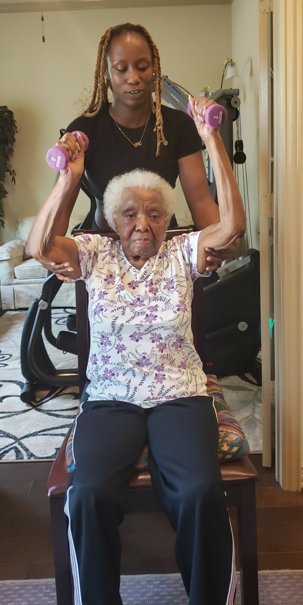 Young woman assisting elderly woman with weights in a living room, both sitting on a chair, woman holding weights overhead.