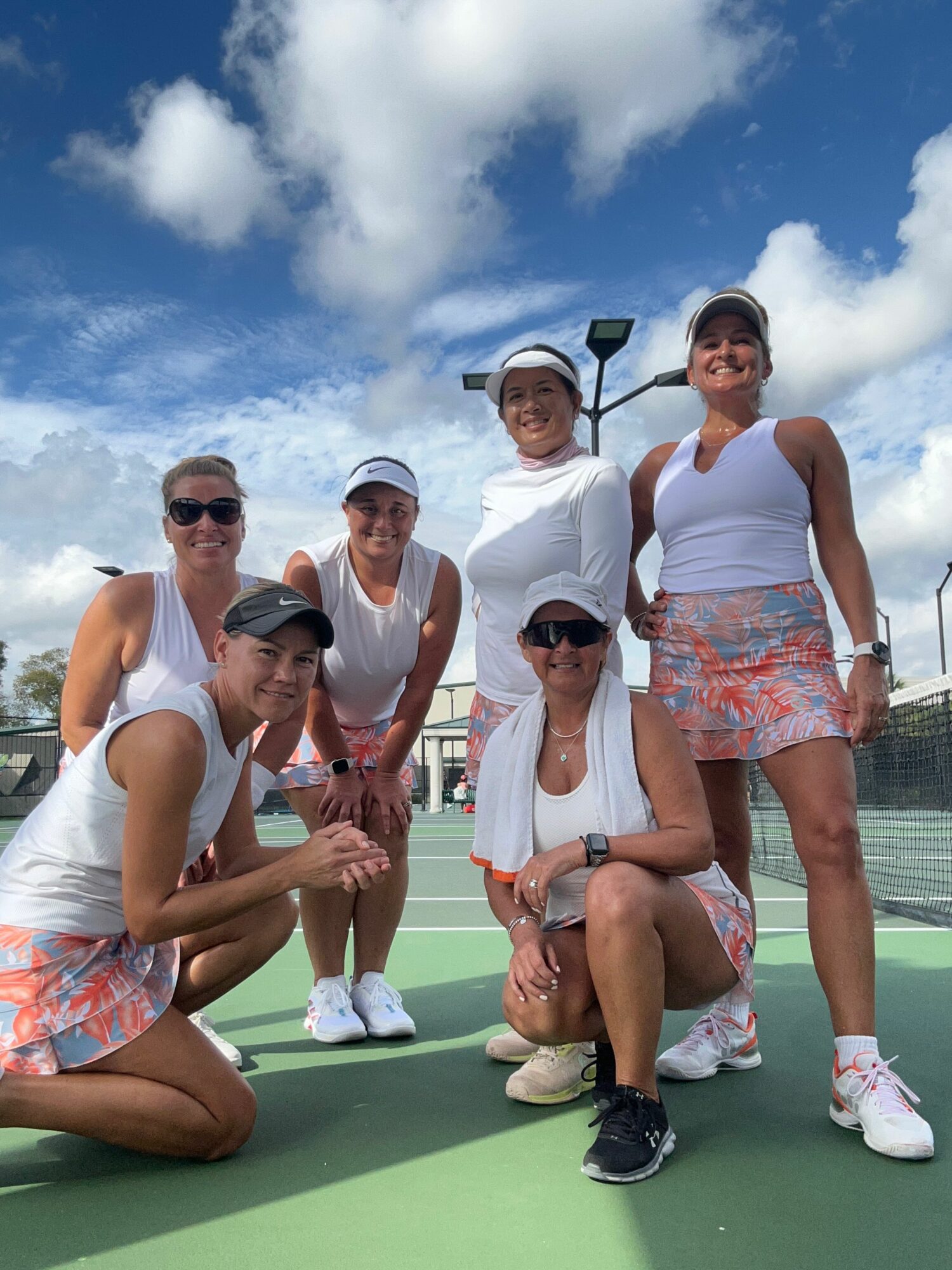 Group of six women on a tennis court, some sitting and some standing, with a blue sky and clouds in background.