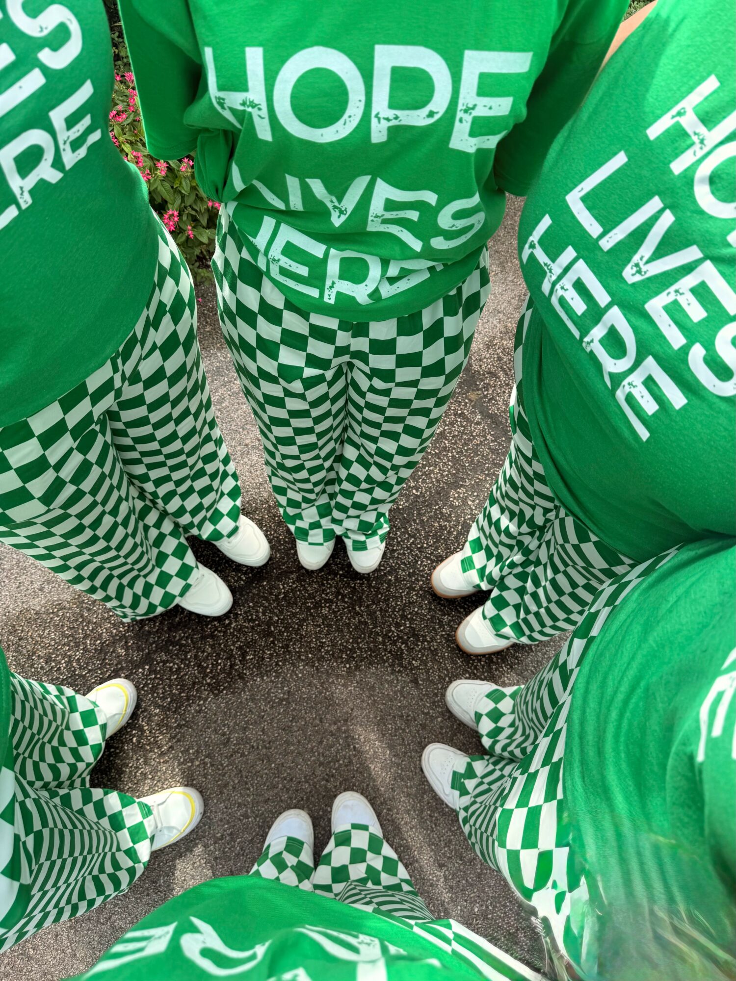 Group of people wearing green shirts with 'HOPE LIVES HERE' and checkered pants, standing in a circle outdoors.