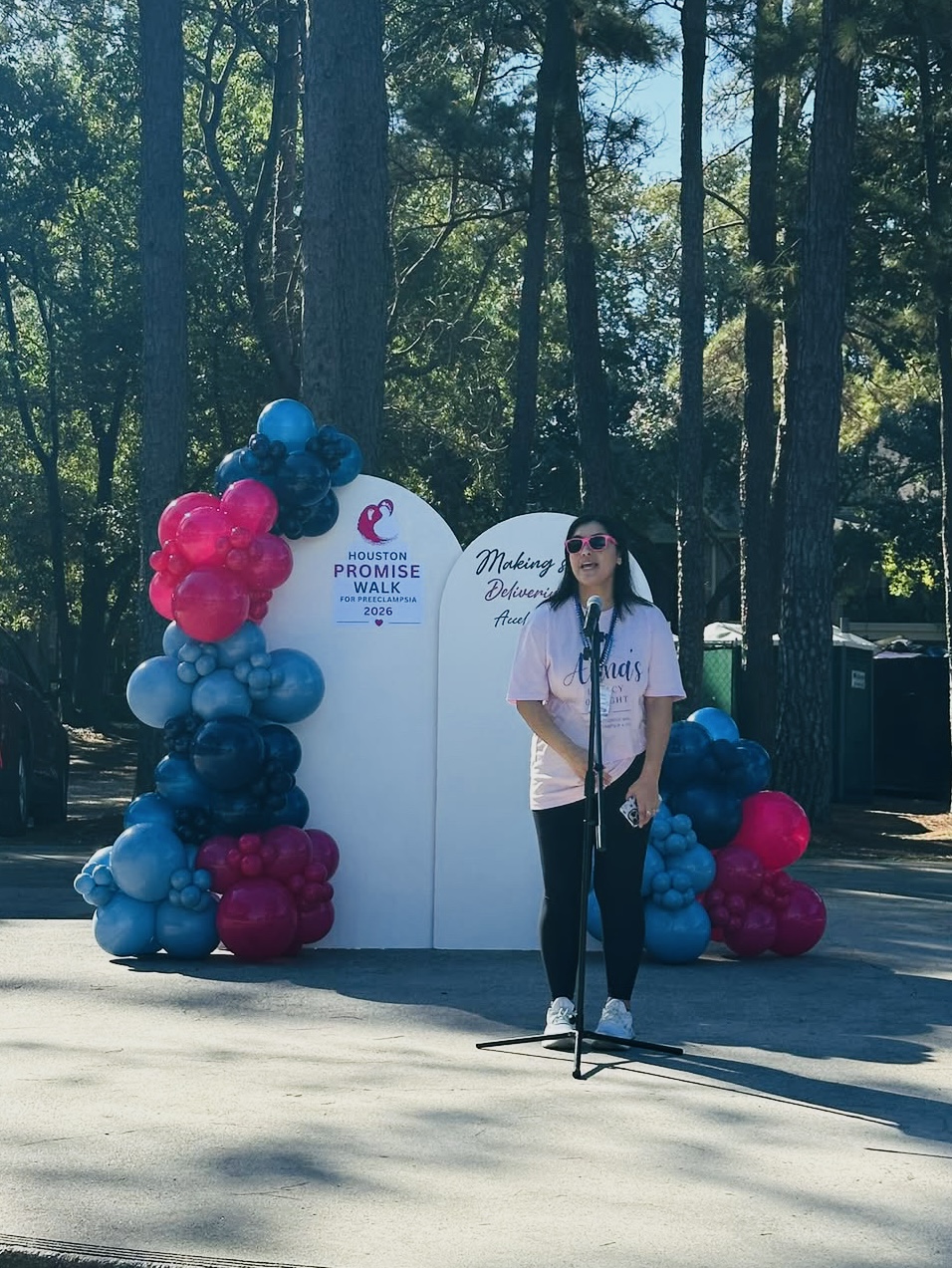 Woman standing outdoors in front of a white backdrop with balloons on each side, trees in the background.