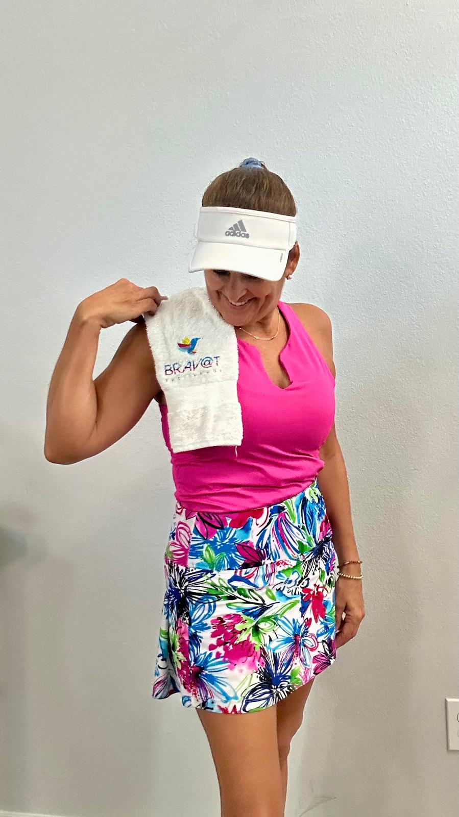Woman in pink top and colorful skirt holding a towel, wearing a white visor, smiling, against a plain background.