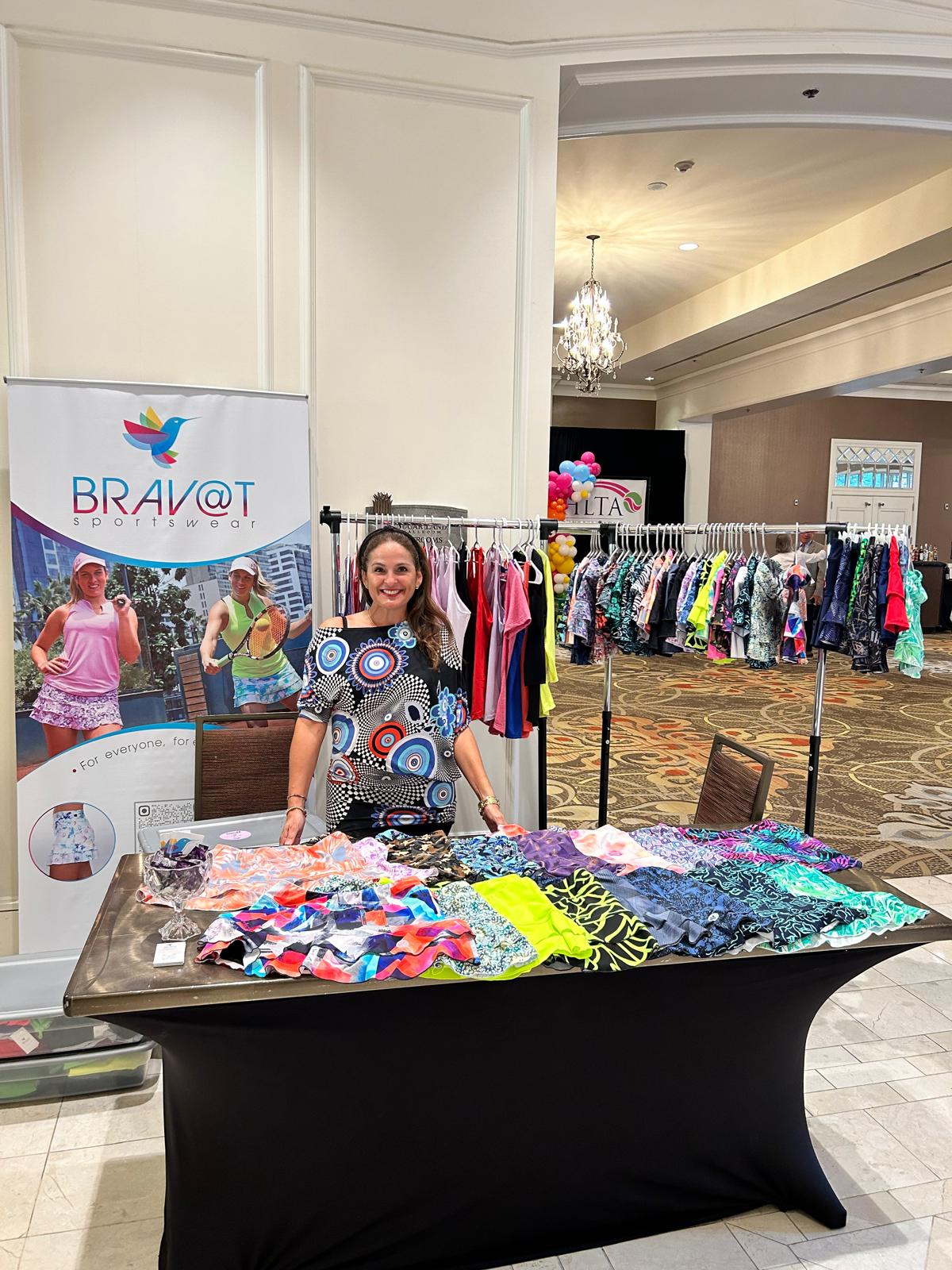 Woman standing behind a table with colorful fabric samples at a clothing event, with racks of clothes in the background.