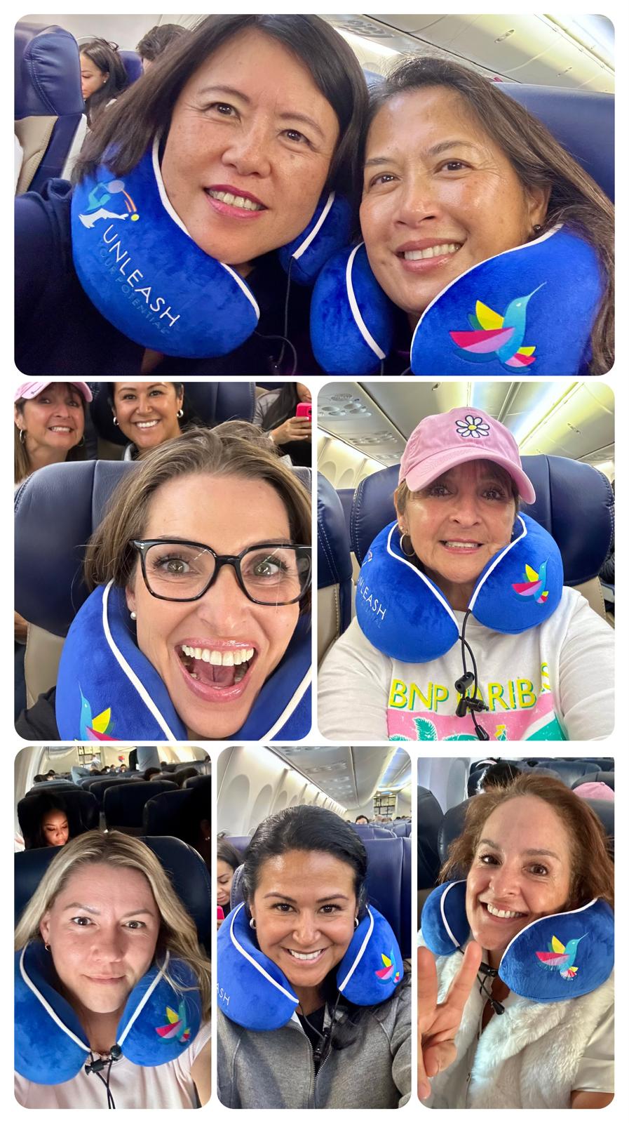 Six women wearing blue neck pillows with colorful designs, smiling inside an airplane cabin.