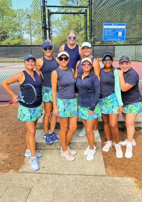 Group of eight women in tennis outfits standing on a tennis court, smiling, with a fence and trees in the background.