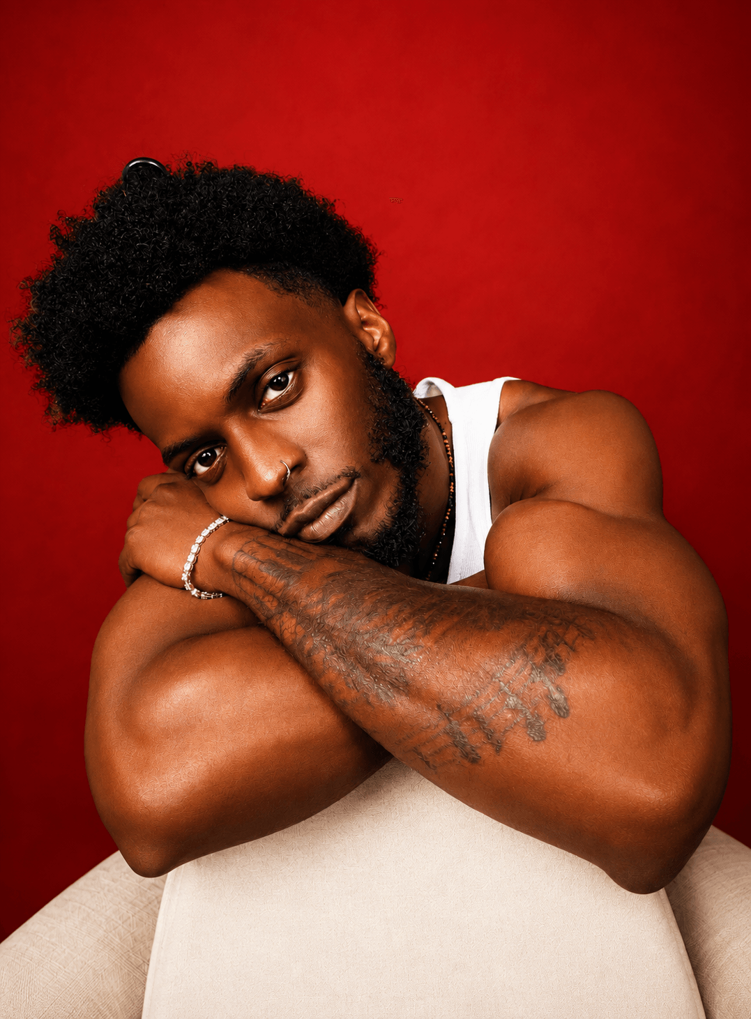 Young man with curly hair and a beard rests his head on crossed arms, wearing a white tank top, against a red background.